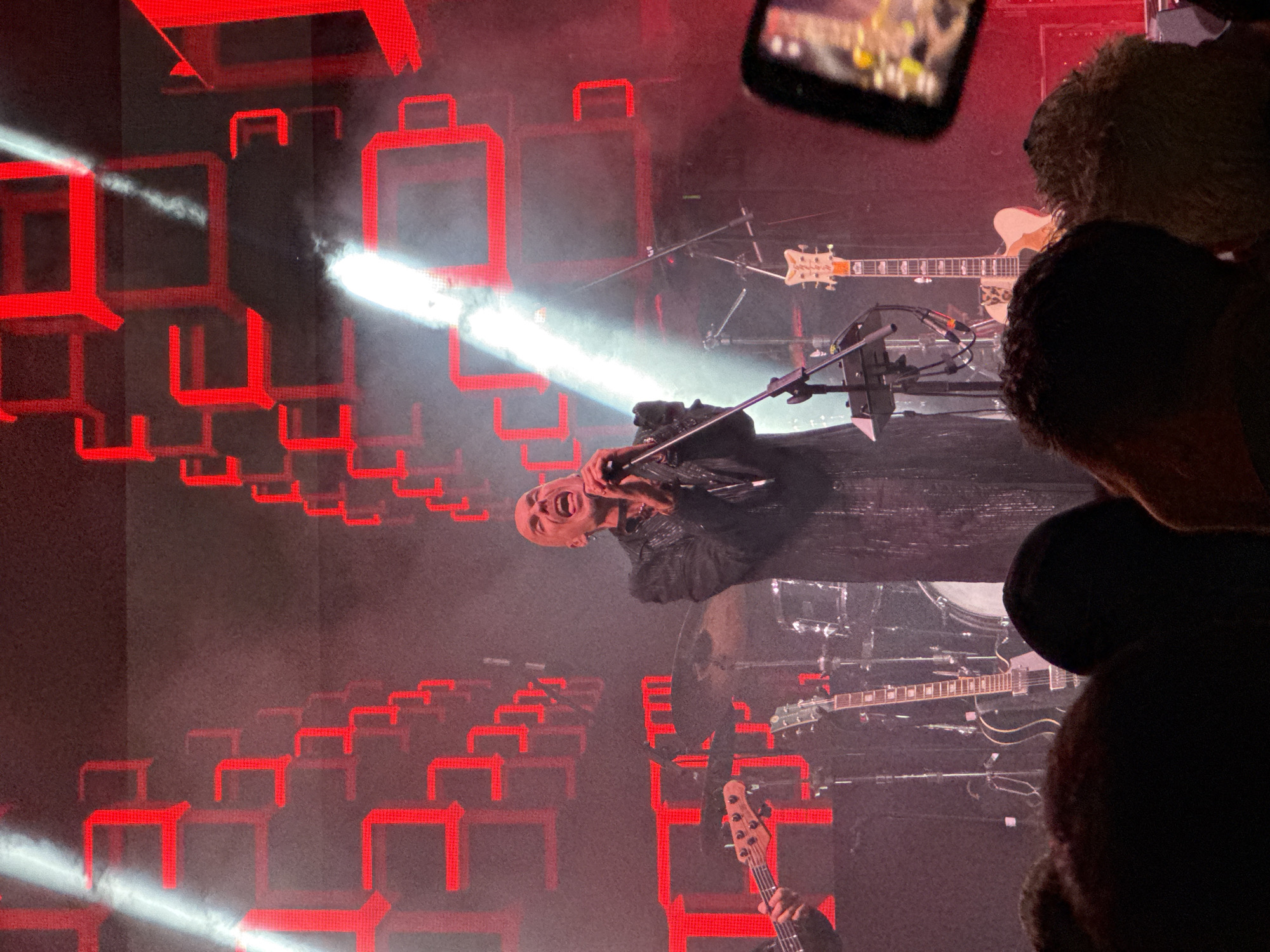 A dynamic and intense photograph captures a live music concert on an indoor stage, likely at night, in Budva, Montenegro. The central focus is a bald male singer, mid-performance, with his mouth wide open in song, holding a microphone to his face. He is wearing a dark, possibly textured or sequined jacket, conveying a passionate and energetic delivery.
The stage is dramatically lit by strong white spotlights that cut through a hazy atmosphere. A striking visual element is a backdrop or screen featuring numerous glowing red rectangular outlines, resembling three-dimensional frames or boxes, which create an abstract and modern visual effect.
Musical instruments are prominent on stage. To the singer's right, a semi-hollow body electric guitar with a distinct headstock and a light-colored, possibly leopard-print body is visible, along with another dark electric guitar and elements of a drum kit. In the foreground, hands can be seen playing a bass guitar. Microphone stands and various cables are also part of the stage setup.
The perspective of the image is from the audience, with several silhouetted heads and shoulders in the foreground, suggesting a close and immersive viewing experience. One audience member in the upper right is actively recording the performance with a smartphone. The overall mood is one of high energy and engagement, driven by the expressive performance and dramatic stage lighting. No discernible text is visible within the frame.