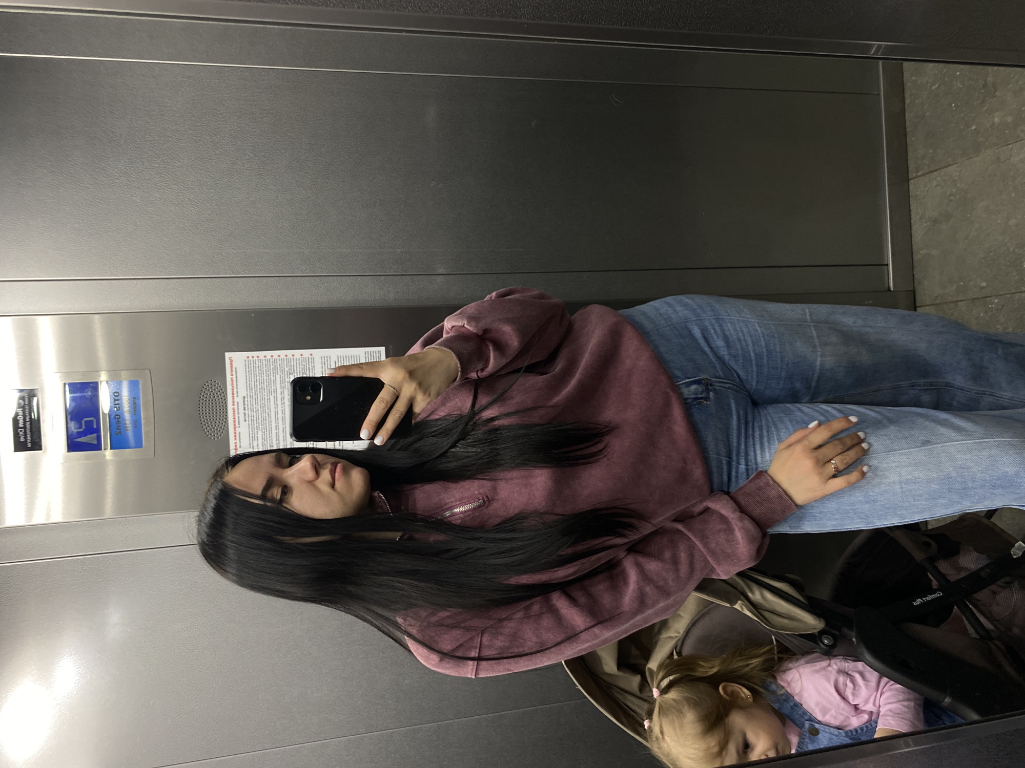 The image depicts a woman taking a selfie in an elevator. The woman has long, dark hair and is wearing a maroon-colored sweatshirt and blue jeans. She is holding a black smartphone. A baby is sitting in a stroller, visible at the bottom of the frame. The elevator has stainless steel walls with signage and indicators. The location is presumed to be in Perm, Russia, based on the prompt.