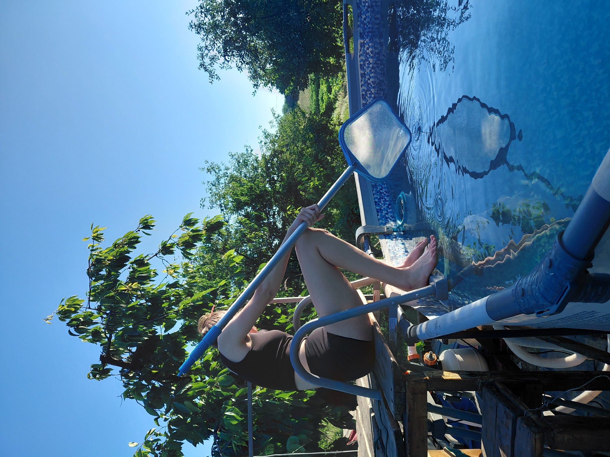 A person is tending to an above-ground swimming pool on a sunny day. The person, wearing a black tank top and black shorts, is positioned on the side of the pool and holding a pool skimmer. They appear to be using the skimmer to clean debris from the water. The pool has a patterned liner visible beneath the clear blue water, and the edge of the pool has a mosaic-like tile pattern.
The setting is outdoors, with lush green trees and foliage surrounding the pool. The sky is a clear, bright blue, suggesting a warm and pleasant day. The sun is high, casting reflections on the water and indicating it is likely midday. The image has a strong sense of summer leisure and outdoor activity. The perspective of the photo is slightly tilted, adding a dynamic feel to the scene. The overall mood is relaxed and focused on the task of pool maintenance. There is no visible text in the image.