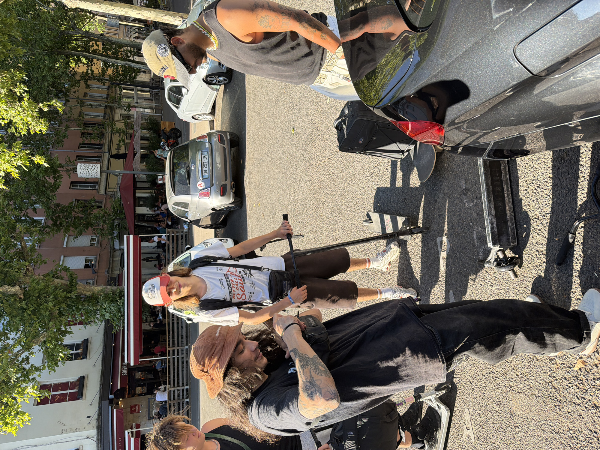 The image captures a group of people gathered on a sunny day in Lyon, France. Two young men are the primary subjects, both wearing casual attire and hats. One man, with long, dark, curly hair, sports a brown corduroy hat and a black t-shirt, his arm adorned with tattoos. The other, wearing a red and white baseball cap and a white t-shirt, has a backpack slung over his shoulder. They appear to be engaged in some sort of gathering or break, possibly after an activity, as there are two scooters visible in the scene.
The background reveals a street with parked cars, one of which is a light-colored Fiat. Buildings with visible window shutters and awnings line the street, suggesting an urban setting, likely a commercial area or residential street. Trees with dense green foliage provide shade, indicating a warm, possibly summer, day. The sunlight casts distinct shadows on the ground, further emphasizing the time of day.
One of the men is holding a long pole or stick, and the presence of the scooters hints at an activity like scootering or a related hobby. The overall mood seems relaxed and casual. There's no explicit indication of strong emotions, but the scene suggests a pleasant, everyday moment. The car license plate visible on the Fiat reads "DV-297-JM." The awnings of a building display the word "SOURIRE," which means "smile" in French, adding a small textual detail to the scene.