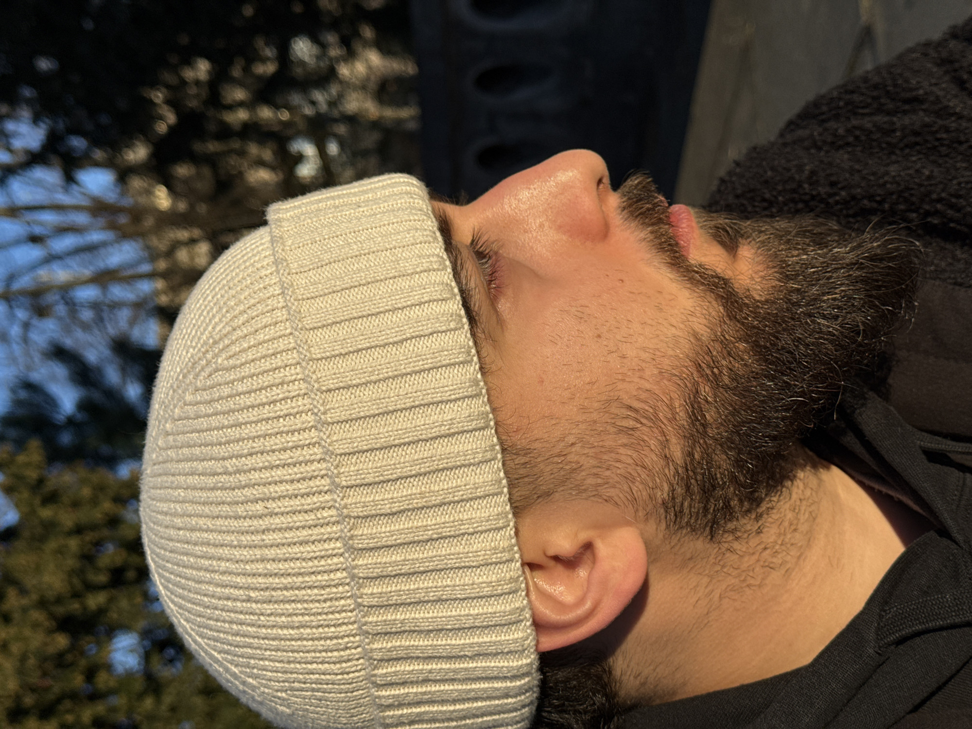 A male individual is depicted in a profile close-up, facing towards the right. He wears a light beige, ribbed knit beanie and has a dark, full beard and mustache. His right ear is visible, and his right eye is partially visible with eyelashes. The skin on his face and neck shows warm illumination from direct sunlight, indicative of golden hour lighting. He is dressed in a dark, possibly black, hooded garment, with a textured, dark outer layer visible over his right shoulder. The background is blurred, featuring green foliage from trees against a blue sky on the left. On the right, a dark stone architectural structure with multiple arched openings is visible. This structure is consistent with the historic Ponts Couverts in Strasbourg, France, specifically elements of its fortified towers or bridge sections overlooking the Ill River.