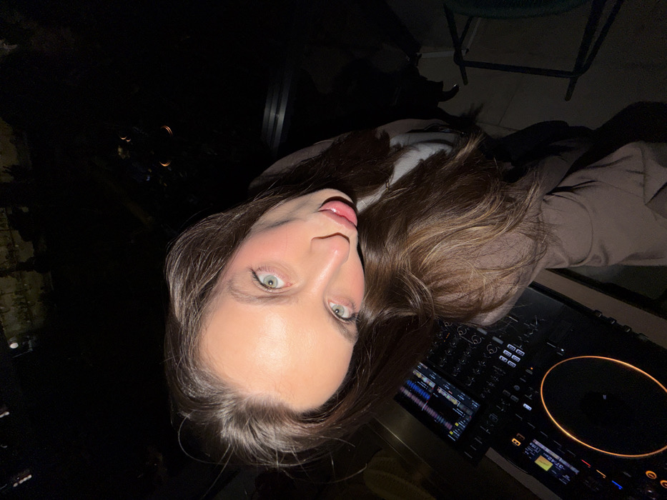 The image displays a female subject in the foreground, looking directly at the camera with her head tilted. She possesses long, dark brown hair and light-colored eyes. Her mouth is slightly open, revealing a portion of her tongue. She is wearing a light-colored top under a brown jacket or coat. To the lower-left, a professional DJ console is visible, featuring an illuminated circular jog wheel, mixer controls, and digital screens displaying track information and waveforms. The setting is dimly lit, suggesting night-time or an indoor venue with ambient lighting. The background outside appears dark with indistinct light sources. A dark chair is partially visible on the right side of the frame. The specific location within the city of Budva, Montenegro, is not discernible from the visual information provided in the image.