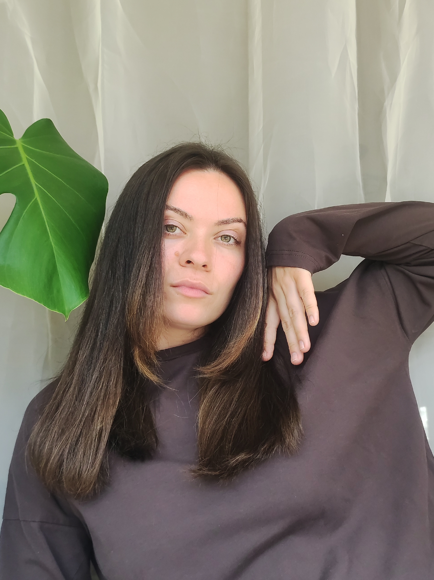 A medium close-up portrait features a woman indoors, located in Chaikovsky, Russia. She has long, straight dark brown hair with subtle lighter streaks, falling over her shoulders and chest. Her light green eyes look directly at the viewer with a calm, neutral expression. She is wearing a dark brown, long-sleeved top. Her right arm is bent, and her hand gently rests on her left shoulder or upper arm. A large, vibrant green Monstera leaf is prominent on the left side of the frame, partially obscuring the background. The background consists of a softly lit, light-colored sheer curtain or fabric with subtle folds. The lighting appears natural, suggesting it is daytime. There is no visible text in the image.