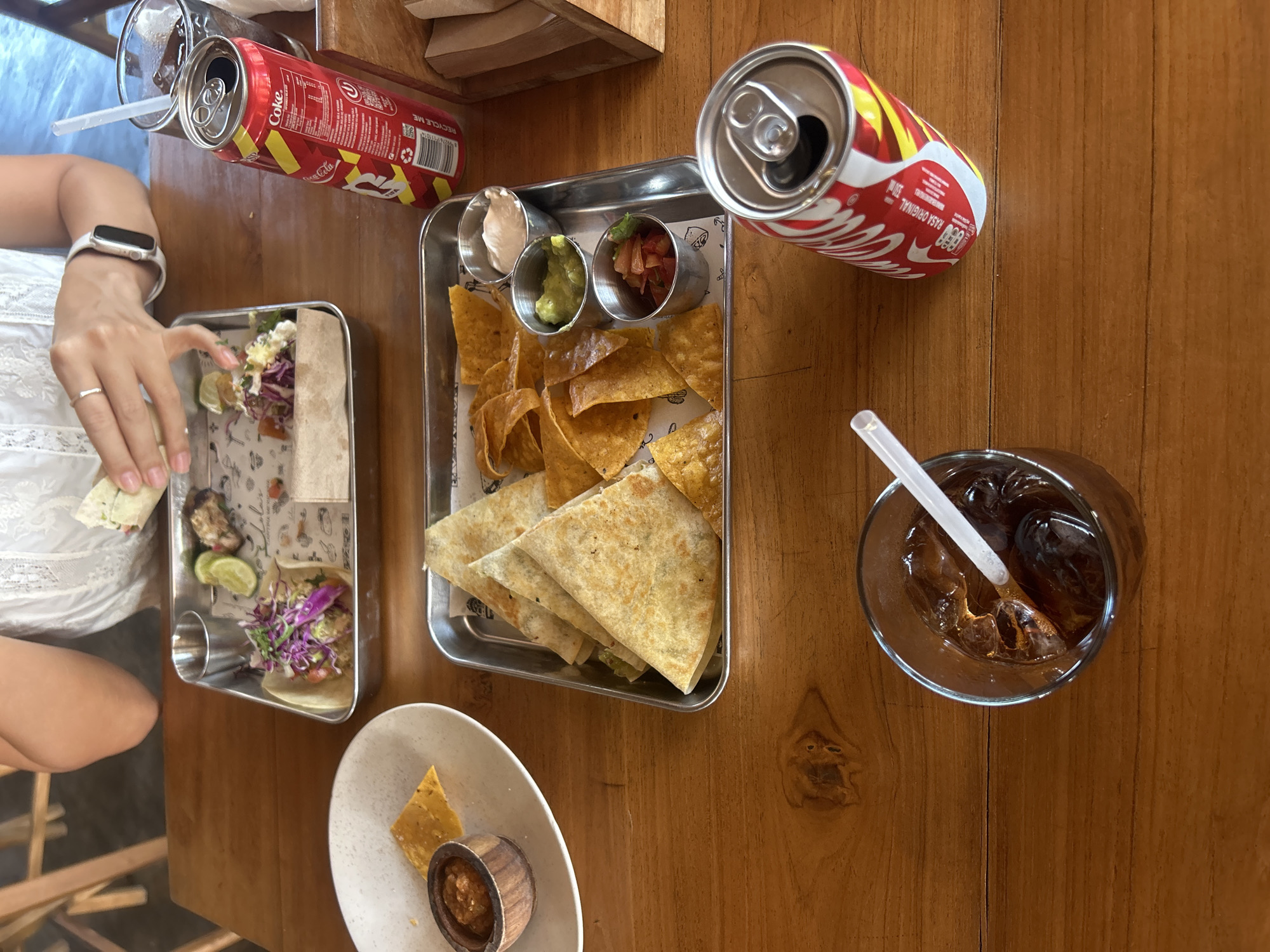 A high-angle shot captures a wooden table laden with food and drinks, likely at a restaurant in Kuta Selatan, Indonesia. In the foreground, a glass of iced cola with a straw sits to the left, while a red and white can of Coca-Cola rests to the right. A metal tray in the middle holds golden tortilla chips alongside three small metal cups containing dips: a white cream, green guacamole, and a red salsa. To the left of this tray, a white plate displays a few tortilla chips and a small wooden bowl filled with salsa. Another metal tray in the upper left quadrant contains a taco with shredded purple cabbage and other toppings, a lime wedge, and a piece of grilled meat, with a person's hands visible, holding a partially eaten taco. The overall scene suggests a casual dining experience.