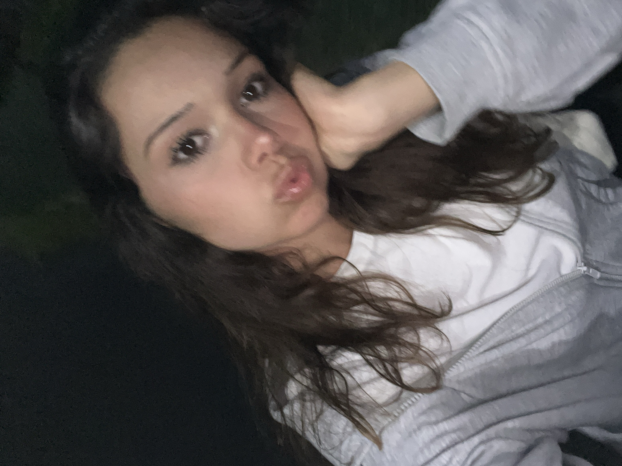 This is a close-up, slightly blurry selfie of a young woman lying down, possibly at night, given the dark background. She has long, dark brown hair that falls around her face and shoulders. Her left hand is resting on her cheek, and her lips are puckered slightly as if blowing a kiss or making a kissing face. She is wearing a white t-shirt underneath a grey zip-up hoodie. The lighting is dim, suggesting an indoor setting or evening outdoors. There is no visible text in the image. The context provided indicates the location is Zlatograd, Bulgaria.
