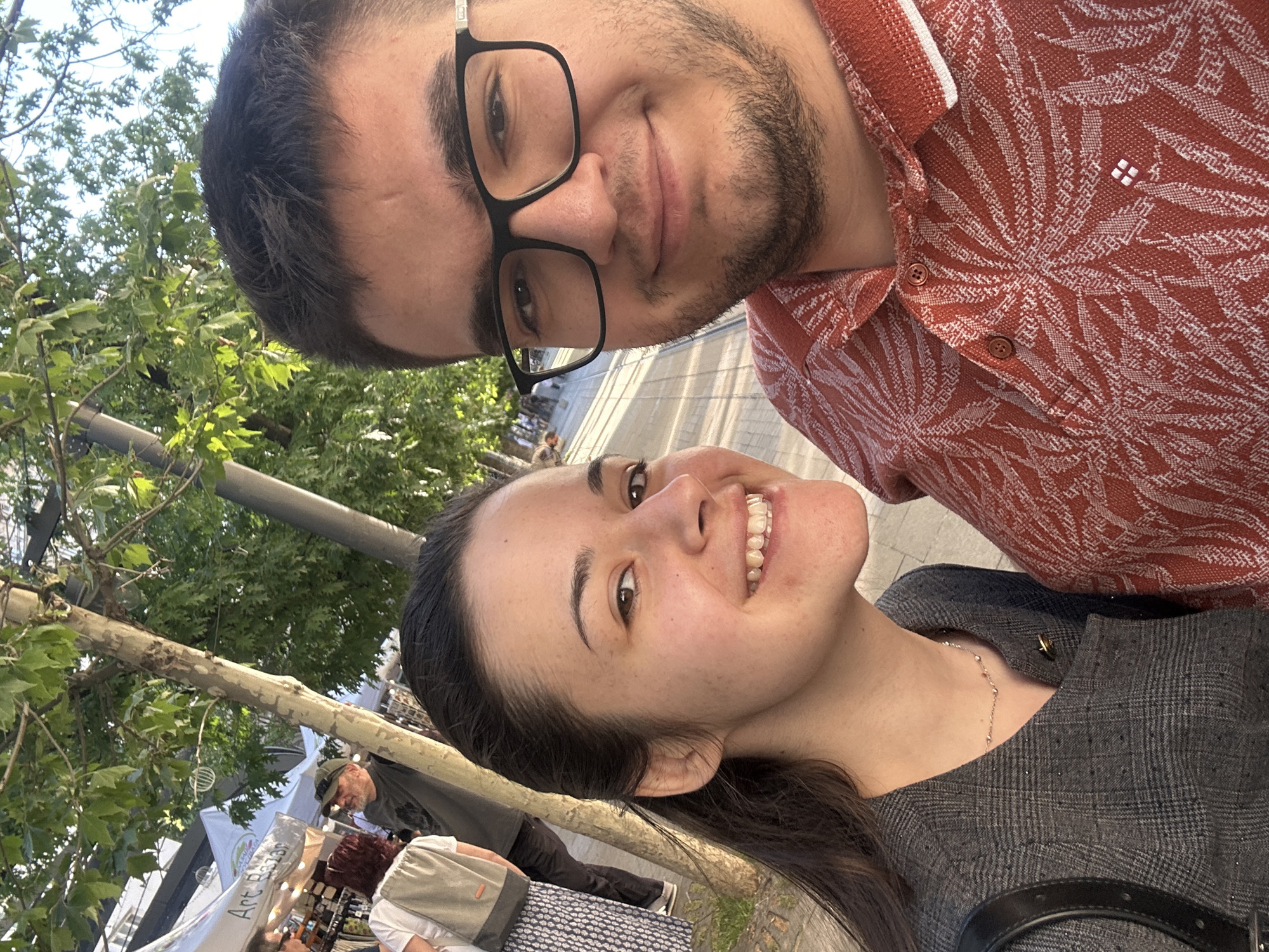 Two individuals are in a selfie, positioned closely together in what appears to be an outdoor public space. The person on the left, closer to the camera, is a woman with dark hair pulled back, a light complexion, and wearing a grey patterned top with a thin silver necklace. She is smiling. The person on the right is a man with dark hair, a short beard, glasses, and wearing a red patterned polo shirt.
The background shows elements of an urban setting with trees, streetlights, and pedestrian traffic, including what appears to be a marketplace or stands with vendors and wares. A sign reading "Art Bazar" is visible in the background. Based on this, the location is likely somewhere in the city of Sofia, Bulgaria.