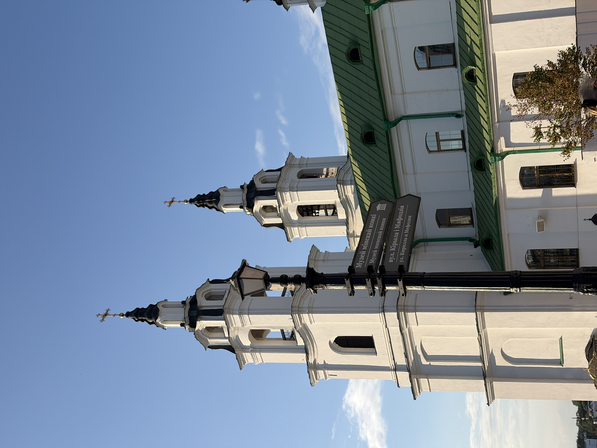 The image shows a white church with two prominent bell towers against a clear blue sky with scattered clouds. One tower is taller than the other and both are topped with black spires and gold crosses. A black lamppost stands in front of the church, with directional signs attached. The signs are in Cyrillic script and indicate locations such as "Museum of Minsk Antiquities" and "St. Cyril and St. Methodius Street". The church has a green metal roof and several windows. A small tree with autumn foliage is visible to the right of the church. The scene appears to be a sunny day in an urban setting.
