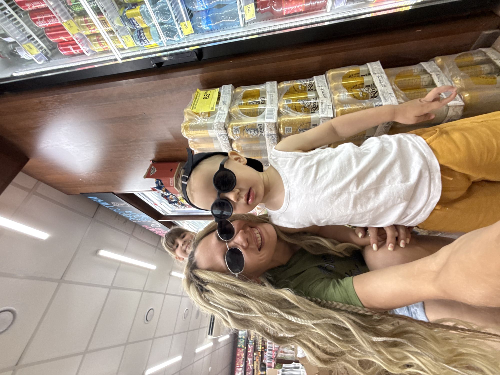 The image captures a candid moment in a grocery store aisle. A woman with long, blonde braided hair and sunglasses is smiling at the camera, holding a young child who is also wearing sunglasses and a white tank top. A second child's head peeks out from behind the woman's shoulder. The background shows shelves stocked with beverages, including cans of beer and bottles of water, typical of a Russian grocery store. The store appears to be in Rusanovo, Russia.