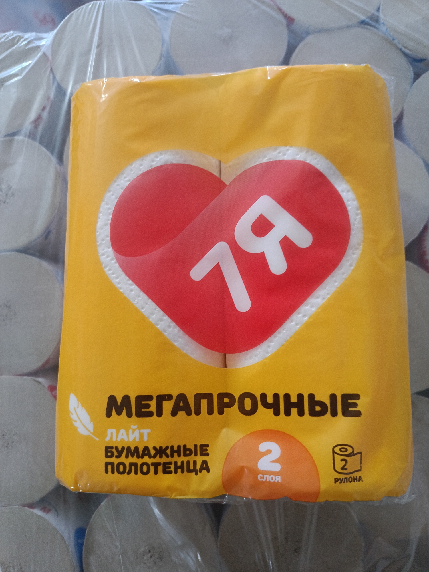 The image shows a close-up of a yellow package of kitchen towels. The package features a large red heart with the number "79" in white inside it. Text on the package is in Russian and reads "МЕГАПРОЧНЫЕ" (Megapročnye, meaning megaproof or ultra-strong), "ЛАЙТ" (Lajt, meaning light), "БУМАЖНЫЕ ПОЛОТЕНЦА" (Bumažnye polotenca, meaning paper towels), "2 СЛОЯ" (2 sloja, meaning 2 layers), and a symbol of a roll with "2 РУЛОНА" (2 rulona, meaning 2 rolls). The packaging is plastic-wrapped and sits in front of a blurred background of stacked white rolls. There are no people visible in the image, and the environment appears to be an indoor setting, possibly a store or warehouse. There is no information in the image to indicate its location in Berdiansk, Ukraine.