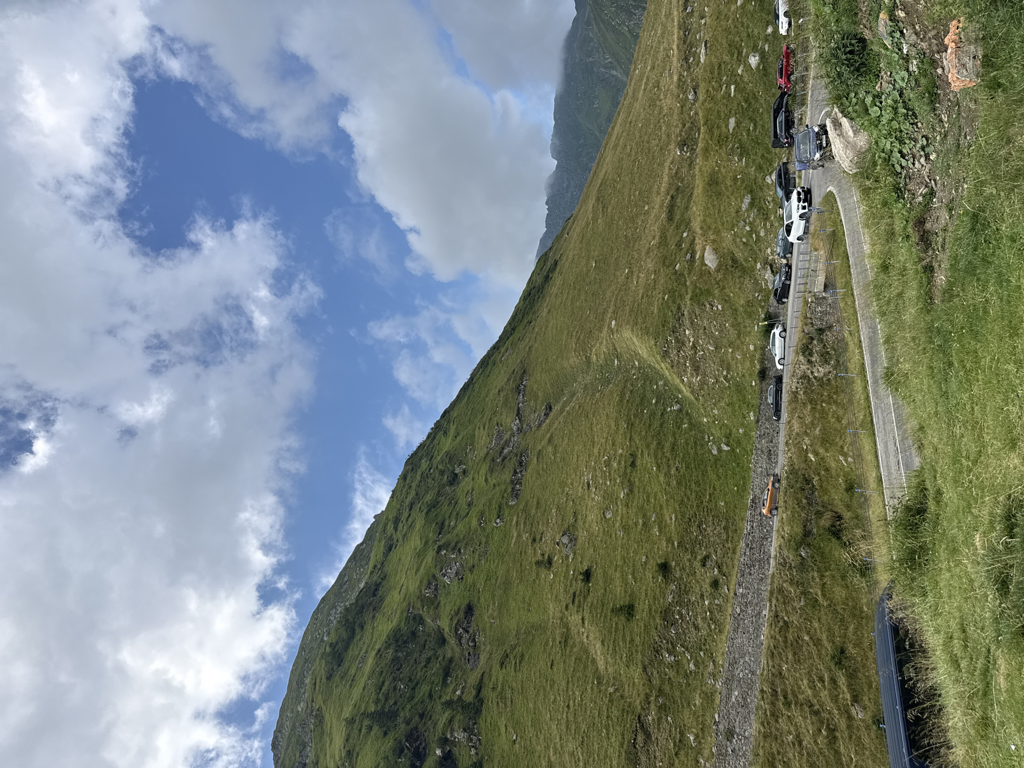 The image captures a scenic view of a winding mountain road in the Transfăgărășan, near Arefu, Romania. The road is bordered by lush green grassy slopes and rocky outcrops. Several cars are parked along the roadside and driving on the road, including a white BMW SUV, a black SUV, a red car, and an orange car. The sky above is partly cloudy with patches of blue. In the distance, the tops of other mountains are visible, some obscured by clouds. The foreground shows overgrown grass and some scattered rocks.