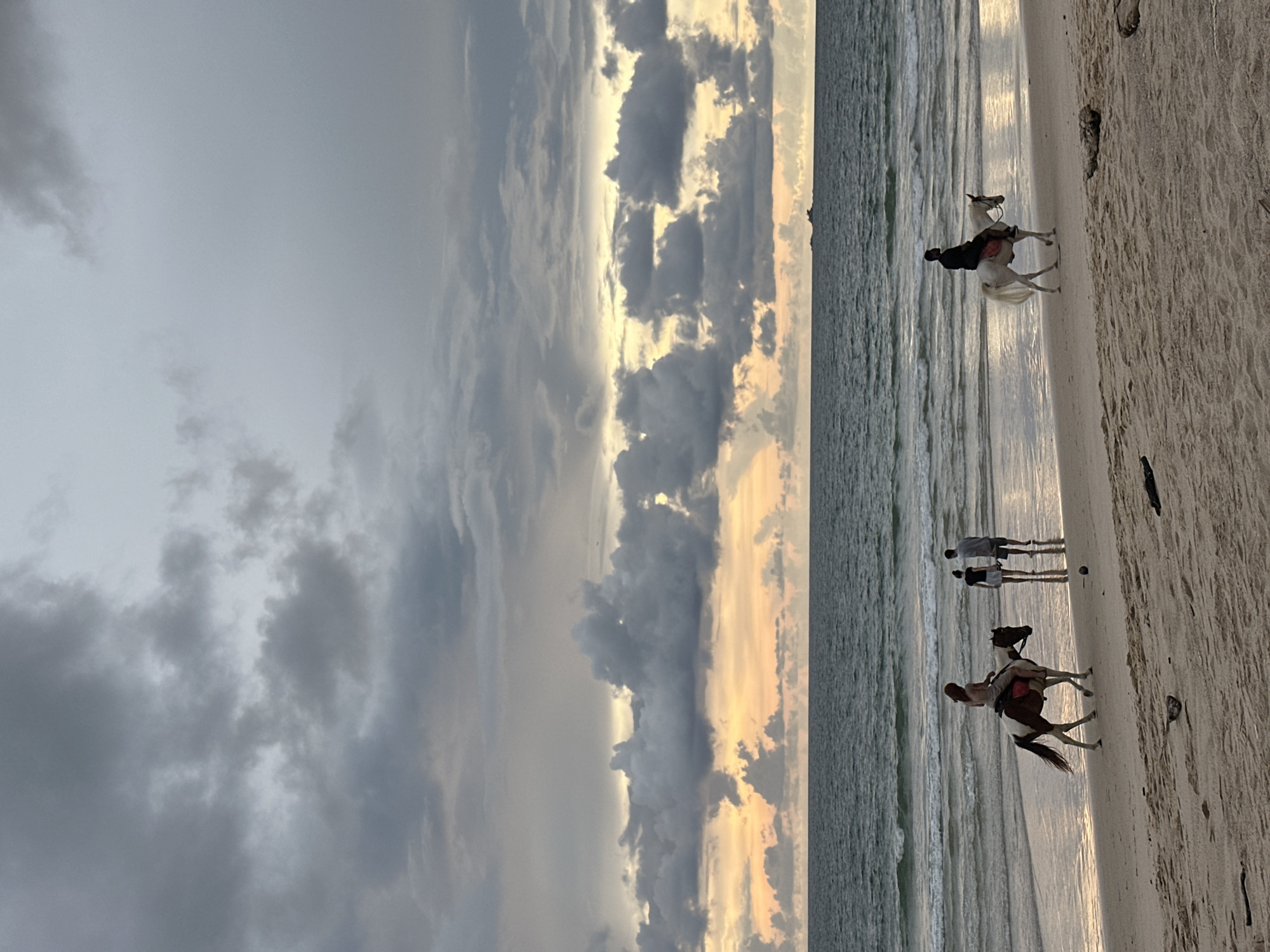 A group of people are on a sandy beach, with a body of water in the background under a cloudy sky. In the foreground, a person rides a brown and white horse from left to right, parallel to the water. To the left of this horse, a couple walks on the wet sand, facing away from the viewer. Further to the right, a person rides a white horse from right to left, also parallel to the water. The sky is filled with heavy, dark grey clouds, with streaks of orange and yellow light breaking through near the horizon. The ocean has moderate waves, and the wet sand reflects the colors of the sky. The overall scene appears to be near sunset.