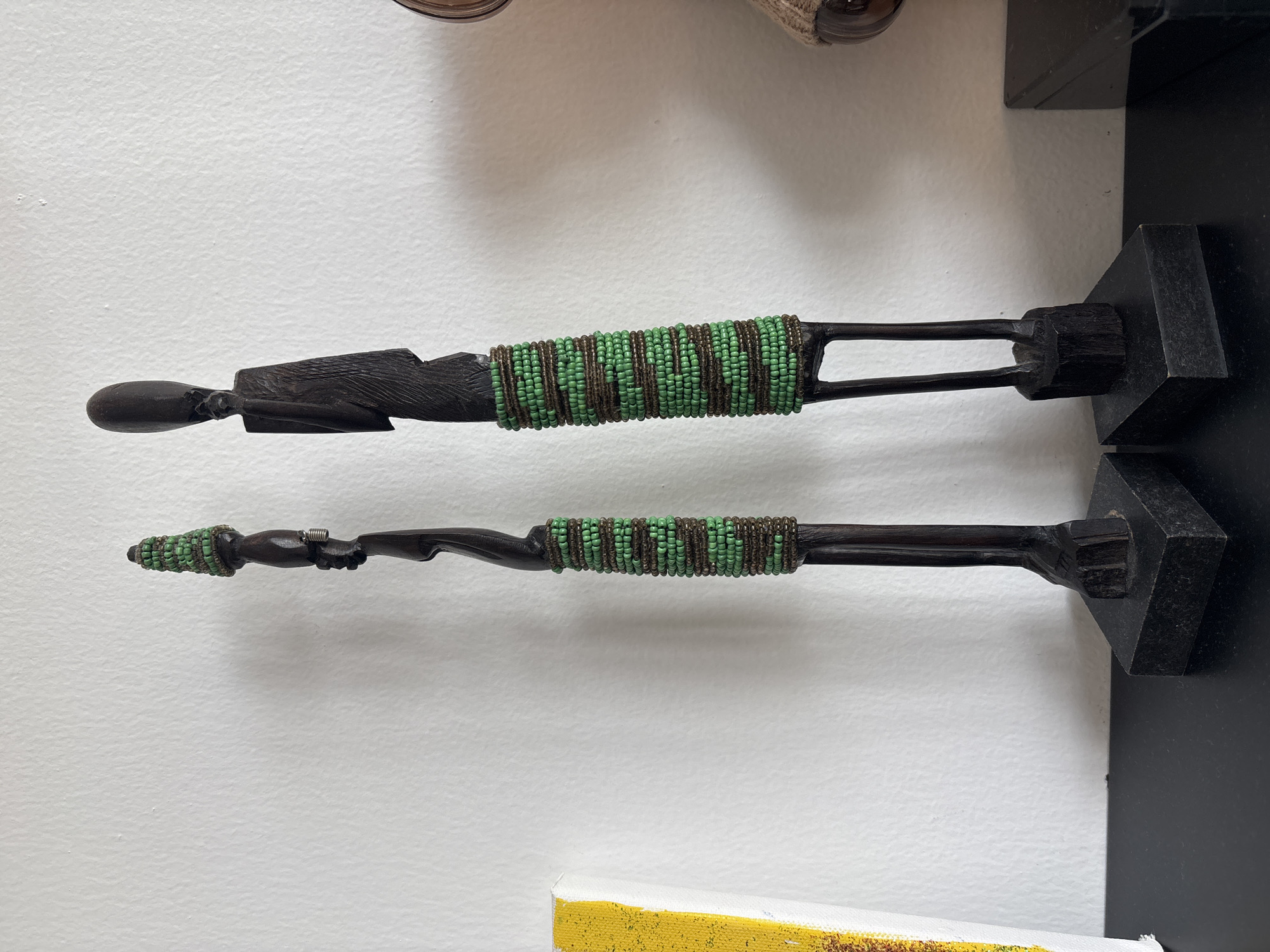 Two dark wooden carved figurines with beaded embellishments stand on a black rectangular base. The figurine on the left is taller and thinner, with a conical head covered in green beads. Its body is adorned with a band of alternating green and brown beads. The figurine on the right is wider, with an elongated head and a garment-like shape around its midsection, also decorated with green and brown beads. Both figures are positioned in a static stance, facing forward. The background is a textured white wall. To the left of the figurines, a portion of a canvas with yellow and white paint strokes is visible. To the right, a hint of a round object and another dark rectangular object are partially in frame. The figurines are placed on a dark surface, likely a shelf or table. The scene is indoors, and no specific location in Dubai, United Arab Emirates can be determined from the provided image.