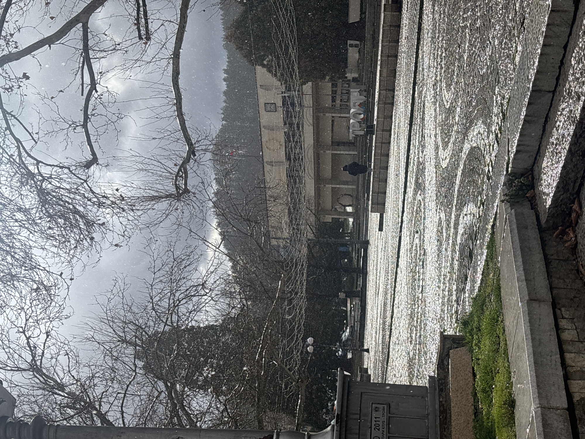 The image depicts a public square in the city of Troyan, Bulgaria, during a light snowfall. In the foreground, a stone monument column is partially visible on the left, bearing engraved text including "2011" and "СТАРИ КЛУБ - ТРОЯН". Adjacent to it, a tiered stone structure contains patches of green grass and scattered brown leaves. The main area of the square is covered by wet, reflective cobblestones laid in a pattern of concentric arcs, shimmering under the light. Bare deciduous trees, some adorned with strings of decorative lights, line the perimeter of the square and extend into the upper frame. A double-headed street lamp stands on the left side of the plaza. A lone individual, dressed in dark attire, walks from left to right across the cobblestone surface, positioned behind a low, concrete barrier. Further back, a prominent white, three-dimensional sign spells "ТРОЯН". The background is dominated by a light-colored, multi-story building with a flat roof, featuring arched ground-floor openings and a flagpole flying at least two flags. Behind this structure, a dense evergreen forest covers a rising hill. The sky is overcast, with visible snow falling across the entire scene, creating bright reflections on the wet surfaces.