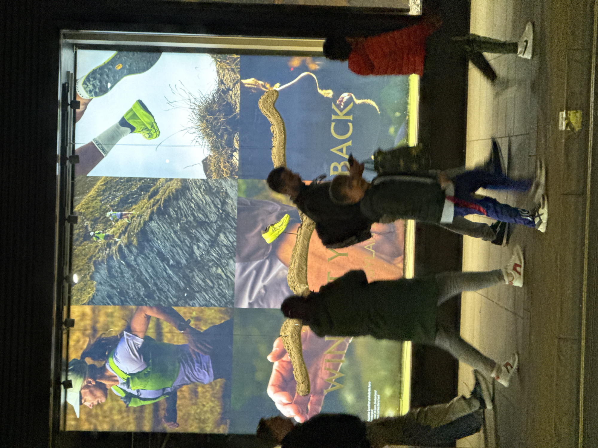 The image depicts a group of pedestrians walking past a storefront display. The display appears to be an advertisement for athletic footwear or outdoor gear, featuring images of shoes and a mountain landscape. The pedestrians, consisting of children and adults, are blurred due to their movement. The scene seems to be set in an urban environment. The reflection on the storefront suggests it is nighttime. Given the pedestrian traffic and storefront display, the location could be a commercial area. Based on the visual style and context, it could be plausibly located in London, United Kingdom.