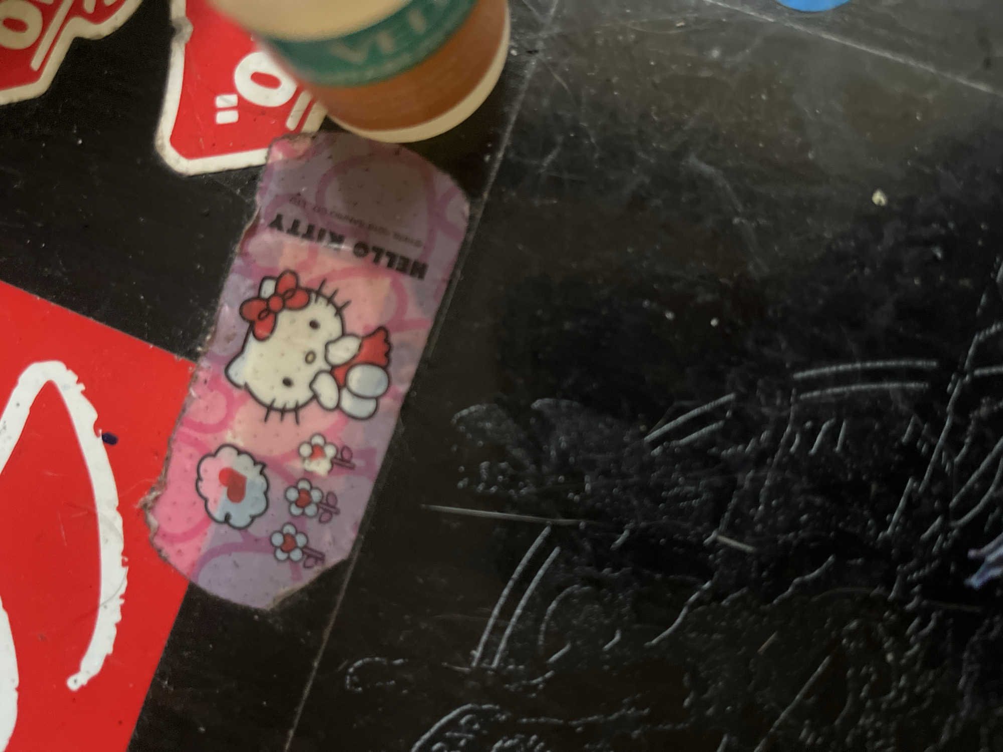 The image shows a close-up, high-angle shot of a black, textured surface, possibly a table or desk. A pink, rectangular bandage with a Hello Kitty design is centered in the frame. The bandage is slightly damaged at the edges and features Hello Kitty with a red bow, surrounded by small flowers and a heart icon. To the right of the bandage, a portion of a spool of thread, primarily brown and green, is visible. In the upper left corner, a red sticker with white lettering is partially in frame, and a red and white sticker with Japanese characters is in the upper right. The background is dark and out of focus.
