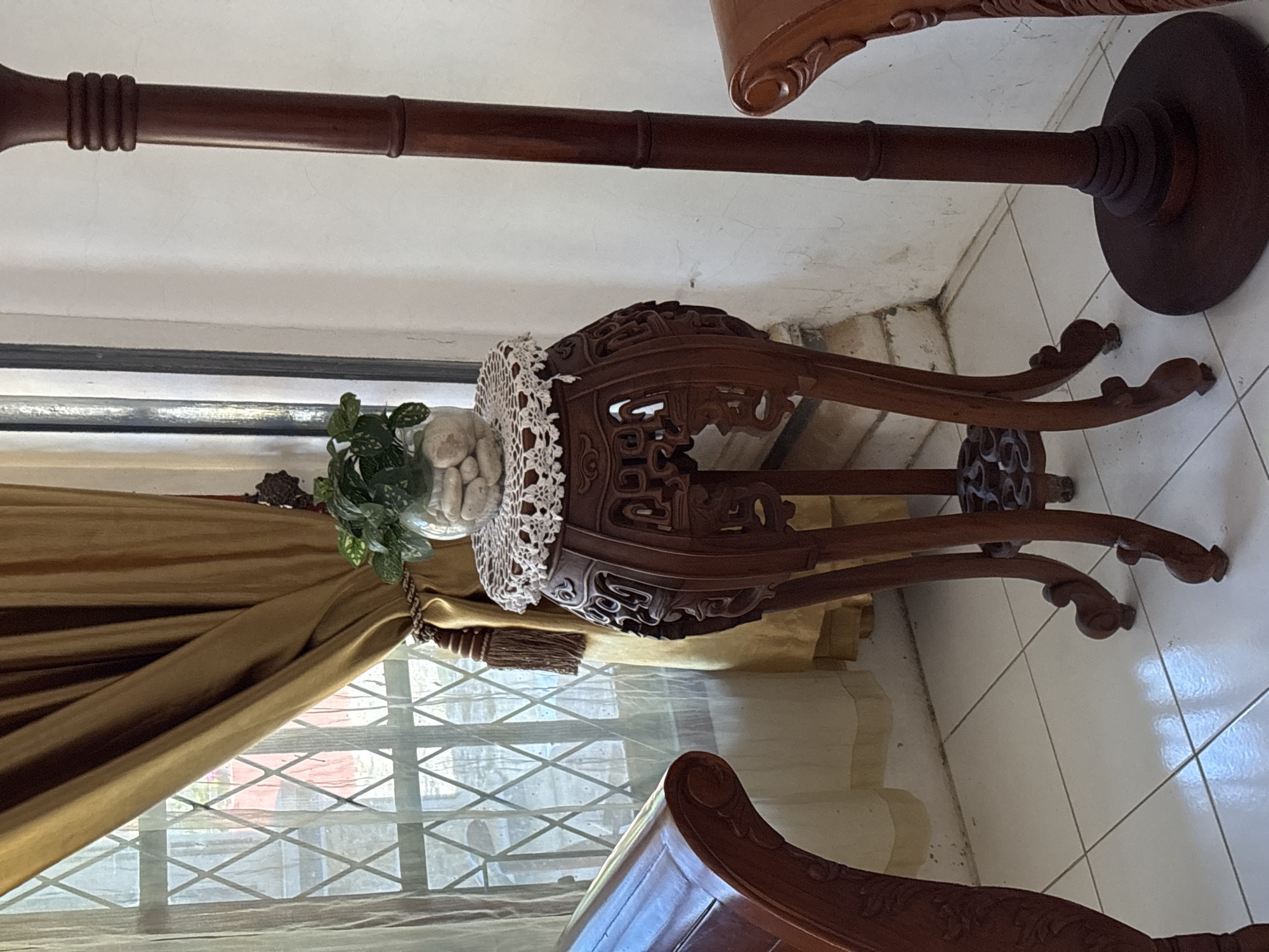 The image shows an indoor scene, likely a living room, with a focus on ornate wooden furniture. A carved wooden pedestal stand is positioned in the center, topped with a glass vase containing a small green plant nestled amongst white stones. The pedestal is draped with a white crocheted doily. To the left of the pedestal, a portion of a curved wooden chair arm is visible. Behind the pedestal, golden curtains hang, partially obscuring a diamond-patterned window. To the right of the pedestal, a tall, dark wooden lamp post stands, with another curved wooden chair arm visible in the far right foreground. The floor is tiled with white square tiles. The overall impression is one of traditional or antique interior decor. The location is identified as Uleepulo, Indonesia.