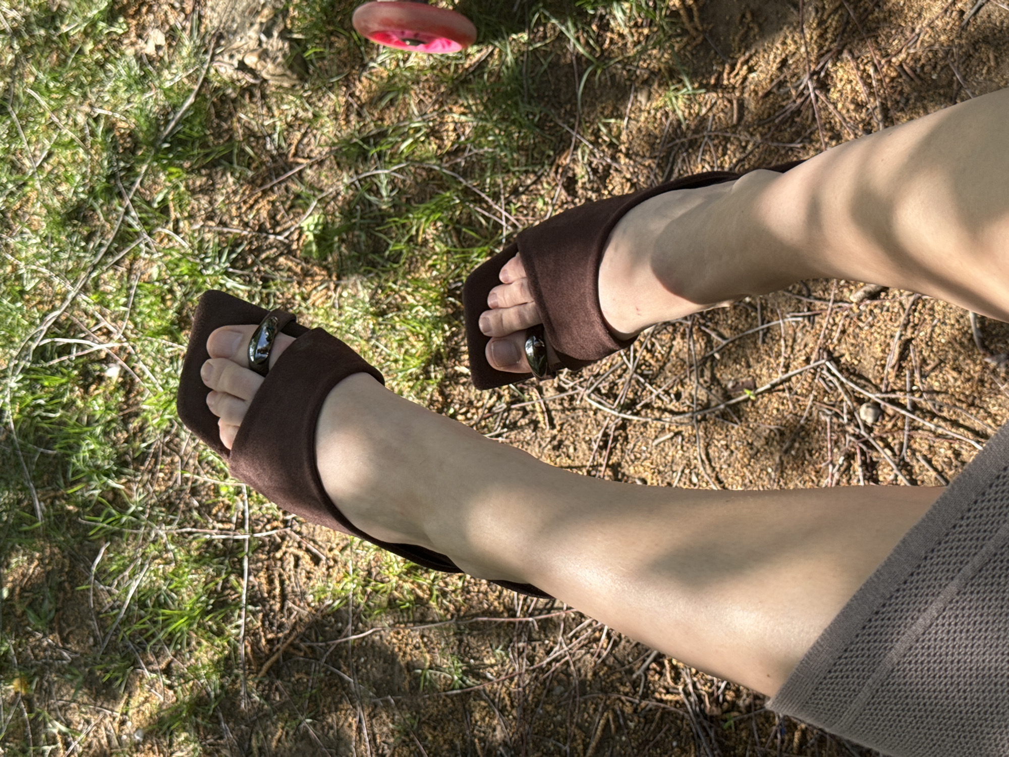 The image depicts a pair of legs and feet wearing brown suede mule-style shoes with toe rings, positioned on a background of grass, twigs, and dry leaves. The perspective is from above, looking down at the legs. A pink object is visible in the upper part of the frame, slightly out of focus. The lower right corner shows the edge of what appears to be a brown knitted garment. The environment suggests an outdoor setting, possibly a park or garden. The location, Sarapul, Russia, cannot be confirmed from the visual information provided.