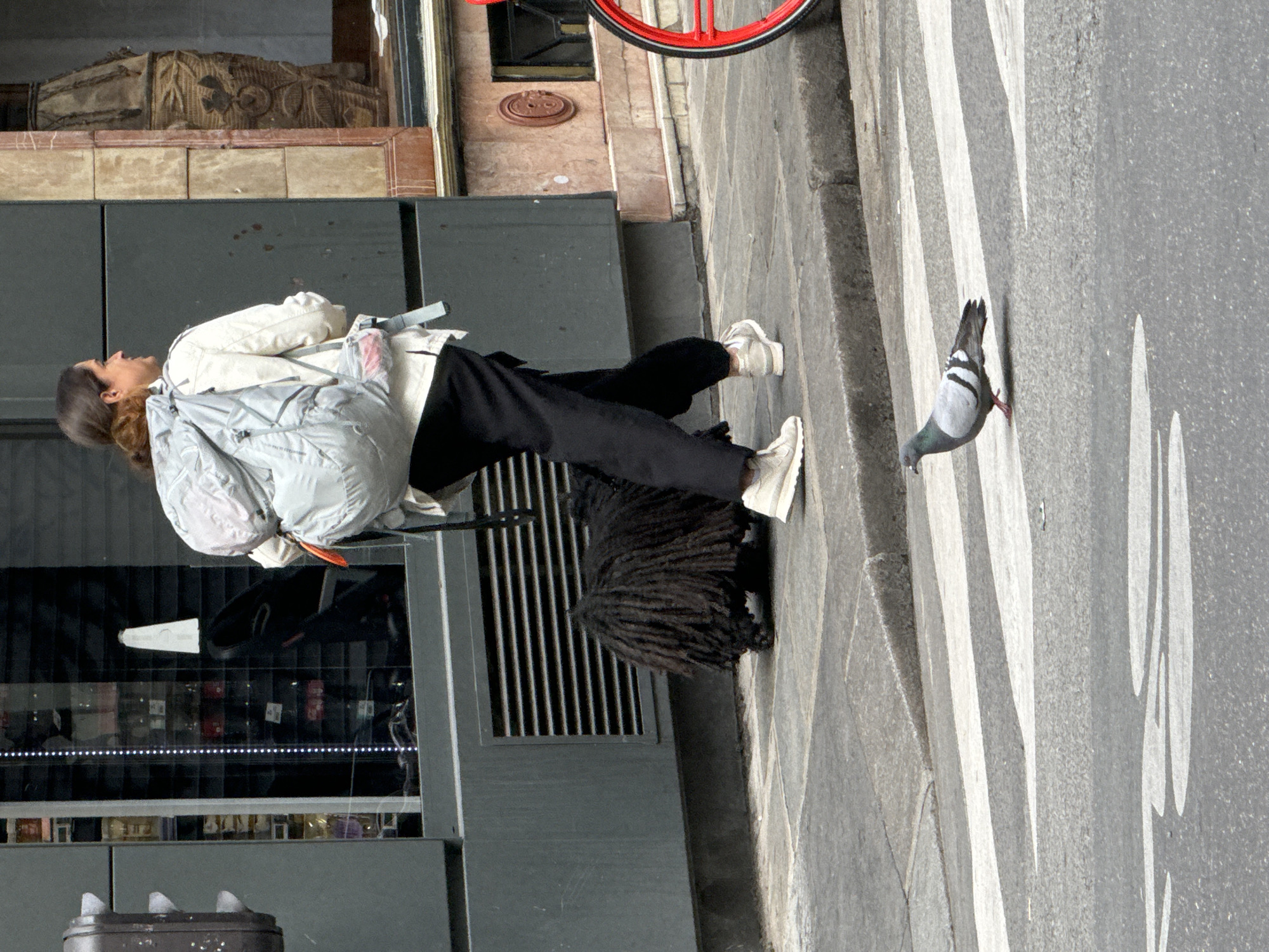 A woman with long brown hair pulled back and wearing a white jacket, black pants, and white sneakers is walking on a city street. She has a light gray backpack. A Komondor dog is beside her. A pigeon is in the crosswalk. The location appears to be Paris, France.