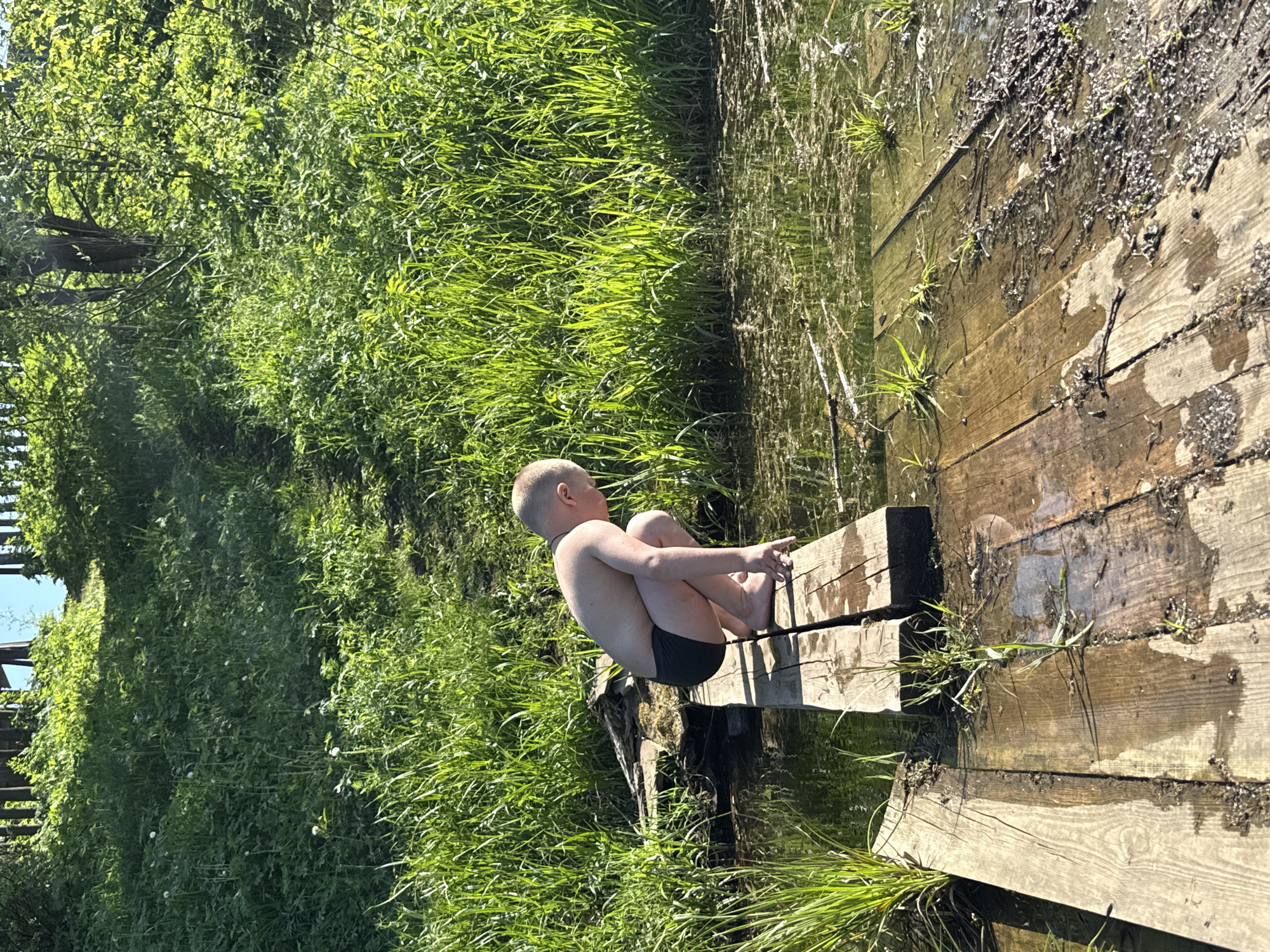 A boy is sitting on a wooden platform by a body of water. The platform is made of wooden planks and appears to be partially submerged. The boy is fair-skinned with short blonde hair, wearing dark swimming trunks. He is in a seated position with his knees bent and his arms wrapped around his legs. The environment consists of green vegetation, including trees and tall grass, alongside the water's edge. This scene is in Zhigalki, Russia.