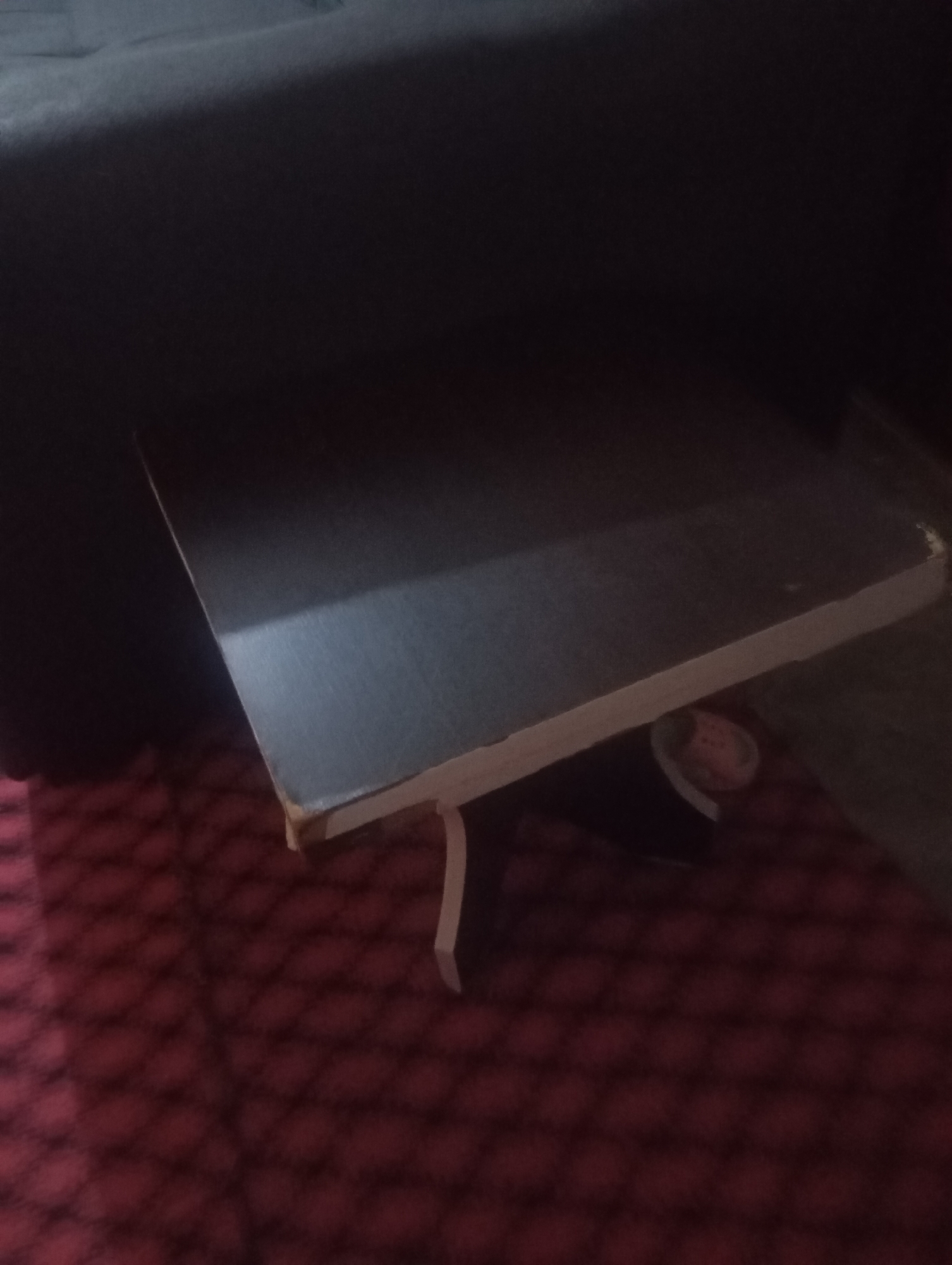 The image shows a dark room with a table and a red patterned carpet. A small, blurry object is visible under the table. The image appears to be a domestic setting. No specific location in Jos, Nigeria, can be determined from the visual information.
