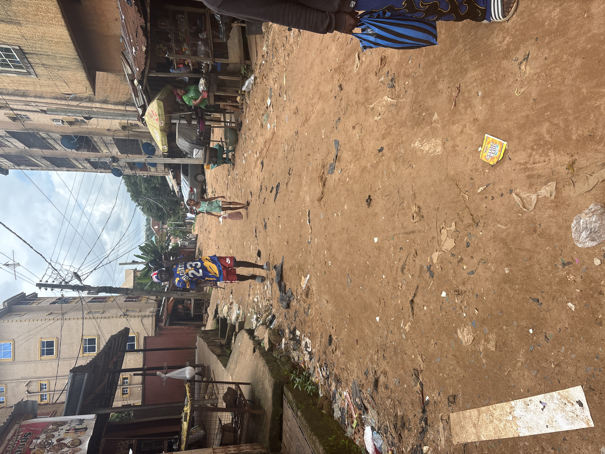 The image shows a street scene in Nkpor, Nigeria. A young boy wearing a blue and yellow basketball jersey with the number 23 is walking down a dirt road. He has a red and white bandana on his head and is carrying a black object over his shoulder. A young girl in a light blue dress is walking in the opposite direction, carrying a shopping bag. In the background, there are multi-story buildings with balconies and satellite dishes. A market stall with a yellow umbrella is visible, and a woman in a green top is attending to customers. The street is littered with trash and debris.
