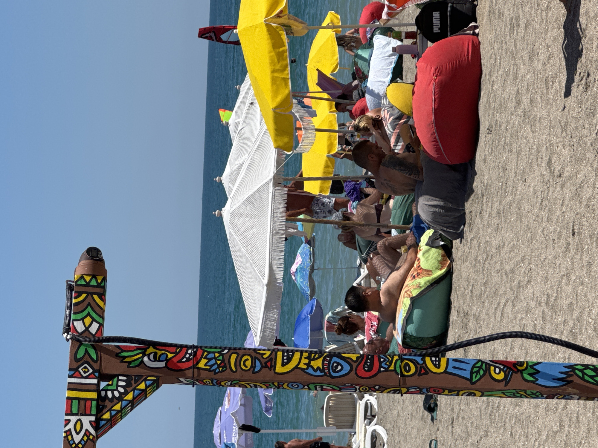 The image depicts a sunny day at a beach in Burgas, Bulgaria, likely near the central beach area, indicated by the presence of numerous beach umbrellas, sunbathers, and the calm sea. In the foreground, a decorative wooden post with colorful, tribal-inspired artwork is prominently featured. A black cable is attached to the post, running vertically.
The beach is populated with people relaxing on beanbag chairs and other seating under various umbrellas, including large white ones and bright yellow ones. The sea is visible in the background, appearing calm and blue. Some individuals are partially submerged in the water. There are also beach chairs and towels scattered across the sand. A red windsurfing sail is visible on the water in the distance, suggesting recreational activities. A black backpack with the "PUMA" logo is placed on the sand near a red beanbag chair. The overall atmosphere is one of leisure and enjoyment of a summer day at the seaside.