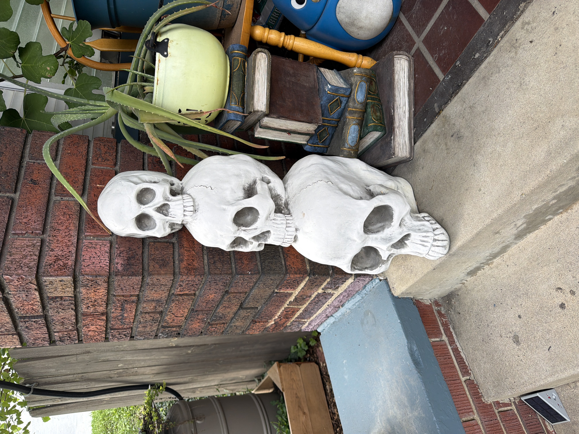 This is a photograph of a stack of three white skull decorations on a concrete porch step. The skulls are stacked vertically, with the largest at the bottom and the smallest at the top. Behind the skulls, a brick column is visible, along with some greenery and a large potted plant. To the right of the skulls, a stack of decorative books and a blue penguin-shaped garden ornament are present. The scene appears to be an outdoor decoration, possibly for Halloween. The location is identified as Pittsburgh, United States of America, although no specific street address or landmark is provided.