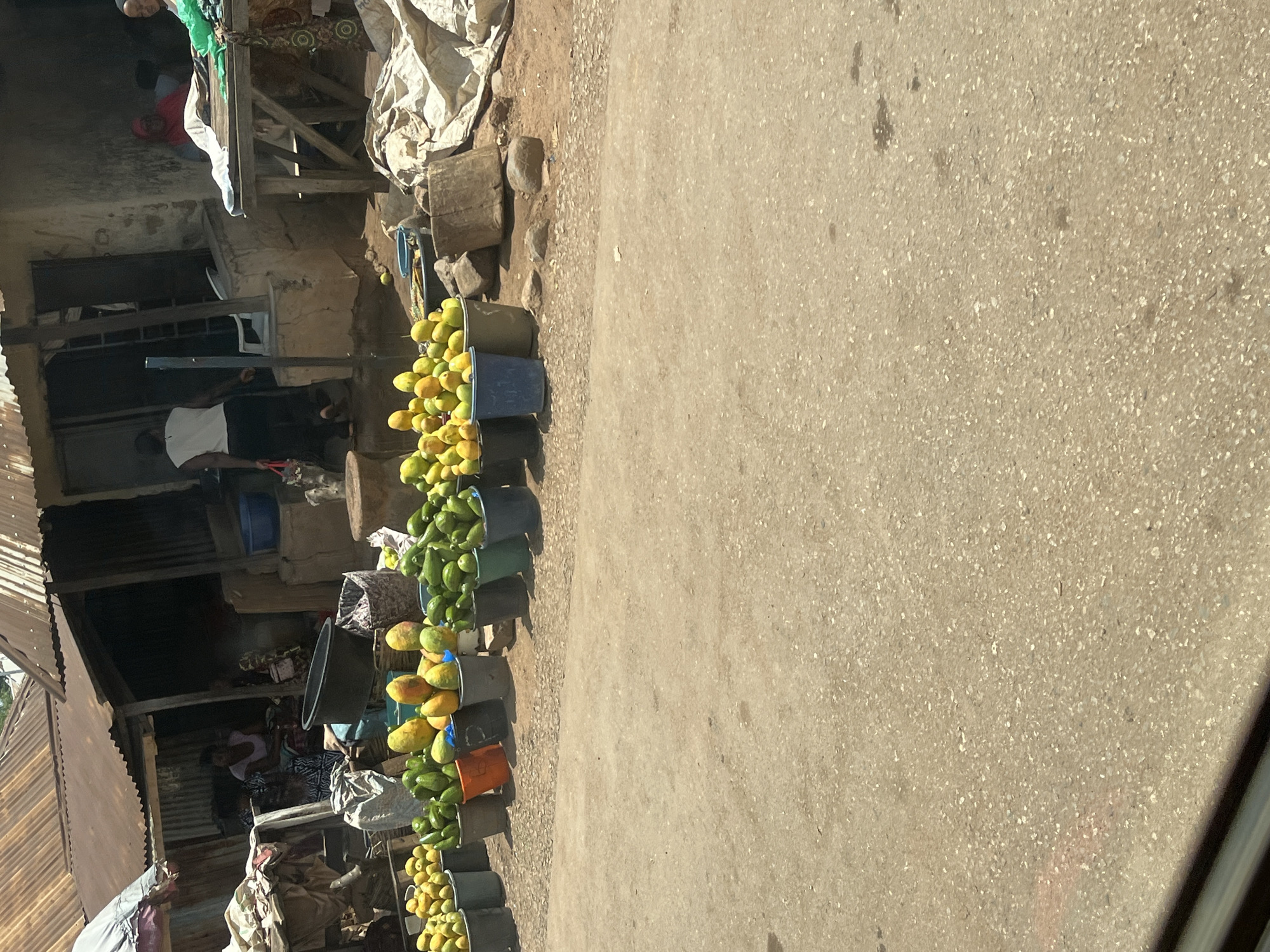 The image depicts a bustling roadside market scene, likely in Karshi, Nigeria, under bright daylight. A long line of plastic buckets, filled with an abundance of mangoes and possibly some green papayas or unripe mangoes, stretches along the edge of a dusty, unpaved road or path. The fruits vary in ripeness, displaying shades of yellow, orange, and green.
On the left side of the frame, behind the fruit display, are several informal market stalls and structures. These are constructed from a mix of corrugated metal sheets, wood, and what appears to be concrete or mud blocks. Tarpaulins are draped over some structures, possibly providing shade or covering goods.
Several individuals are visible within the market. One person wearing a white shirt and dark trousers stands near the fruit buckets, while another in a red top can be seen further back. Inside one of the stalls, a person in a patterned dress is seated, suggesting a vendor attending to their wares. The overall atmosphere is that of a lively, functional outdoor market.
The ground is a light brown, speckled surface, indicative of a mix of dirt and gravel. Shadows are sharp and distinct, confirming a bright, sunny day. No discernible text is visible in the image.