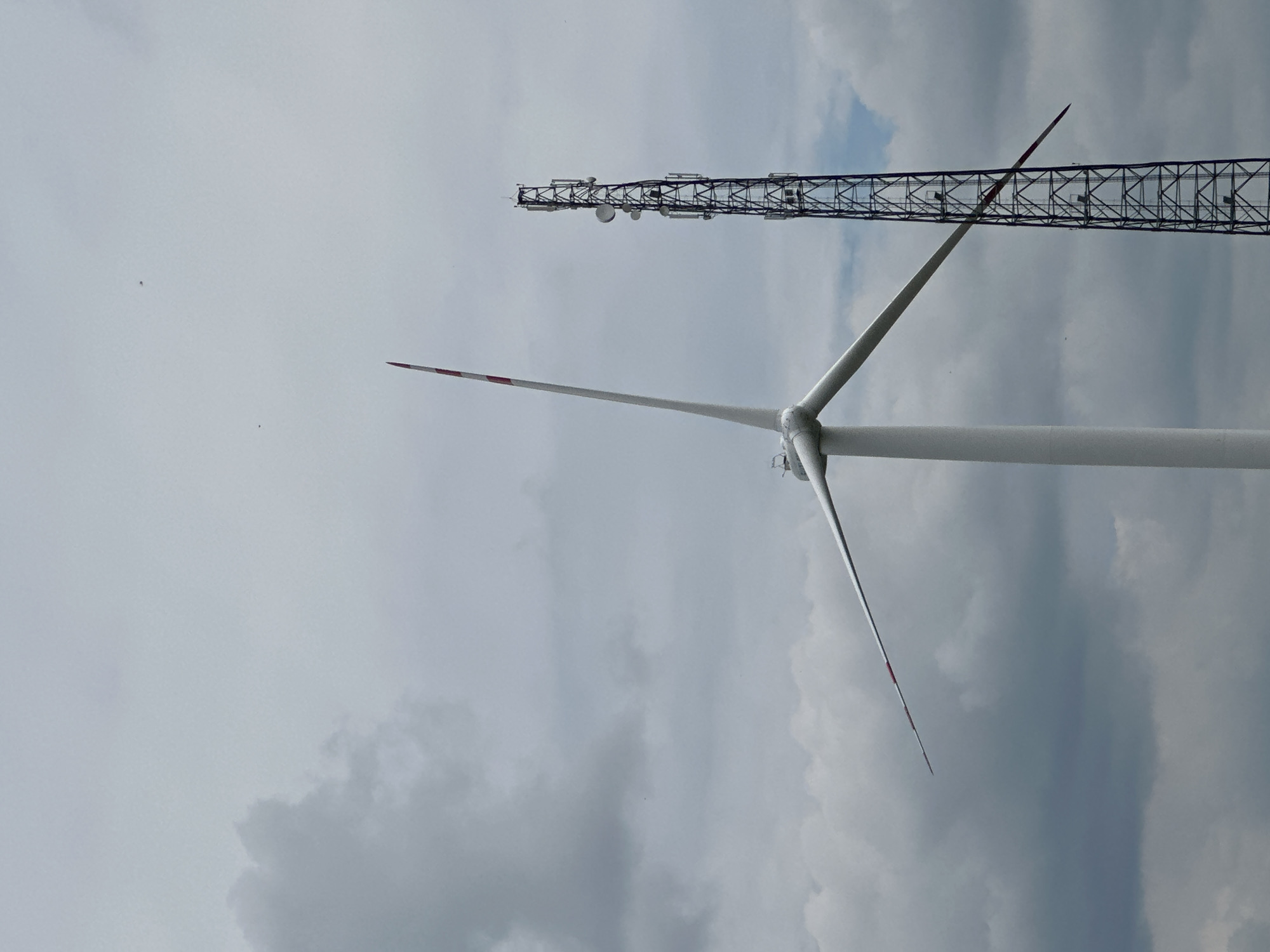 A low-angle, full shot captures a tall, white wind turbine with a single blade positioned diagonally to the right, and a dark, lattice-style telecommunications tower to its right. The sky is overcast with varying shades of gray clouds, with lighter patches and some darker formations. The wind turbine's blade, painted with red and white stripes near its tip, extends across the frame. The telecommunications tower is also dark and appears to have multiple antennas or dishes mounted on its structure at different heights. Two small, dark specks, possibly birds or insects, are visible in the sky. The scene is set against a muted, cloudy backdrop.