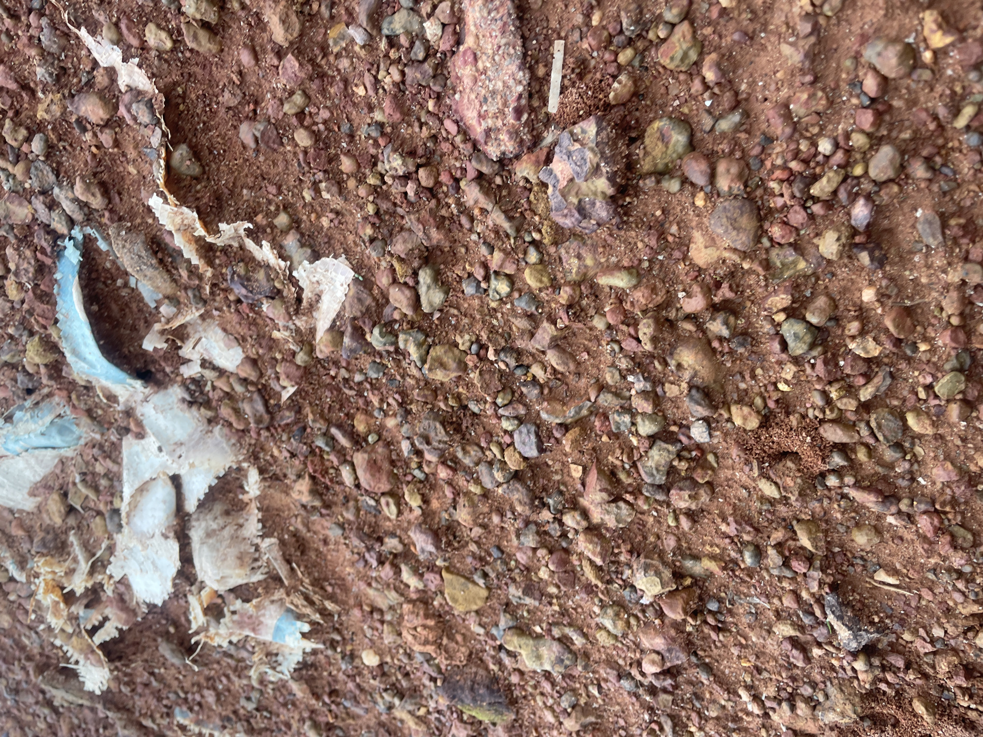 The image displays a close-up, ground-level view of reddish-brown soil interspersed with small pebbles and gravel. Scattered across the surface are fragments of white and light blue material, appearing to be pieces of plastic sheeting or packaging, some of which are torn and weathered. The textures suggest an outdoor environment, likely a disturbed ground area. This scene is consistent with litter found on the roadside or in unkempt areas within the city of Jos, Nigeria. No people are visible in the image, and there are no discernible actions or interactions taking place. The overall impression is one of a mundane, debris-strewn ground surface.