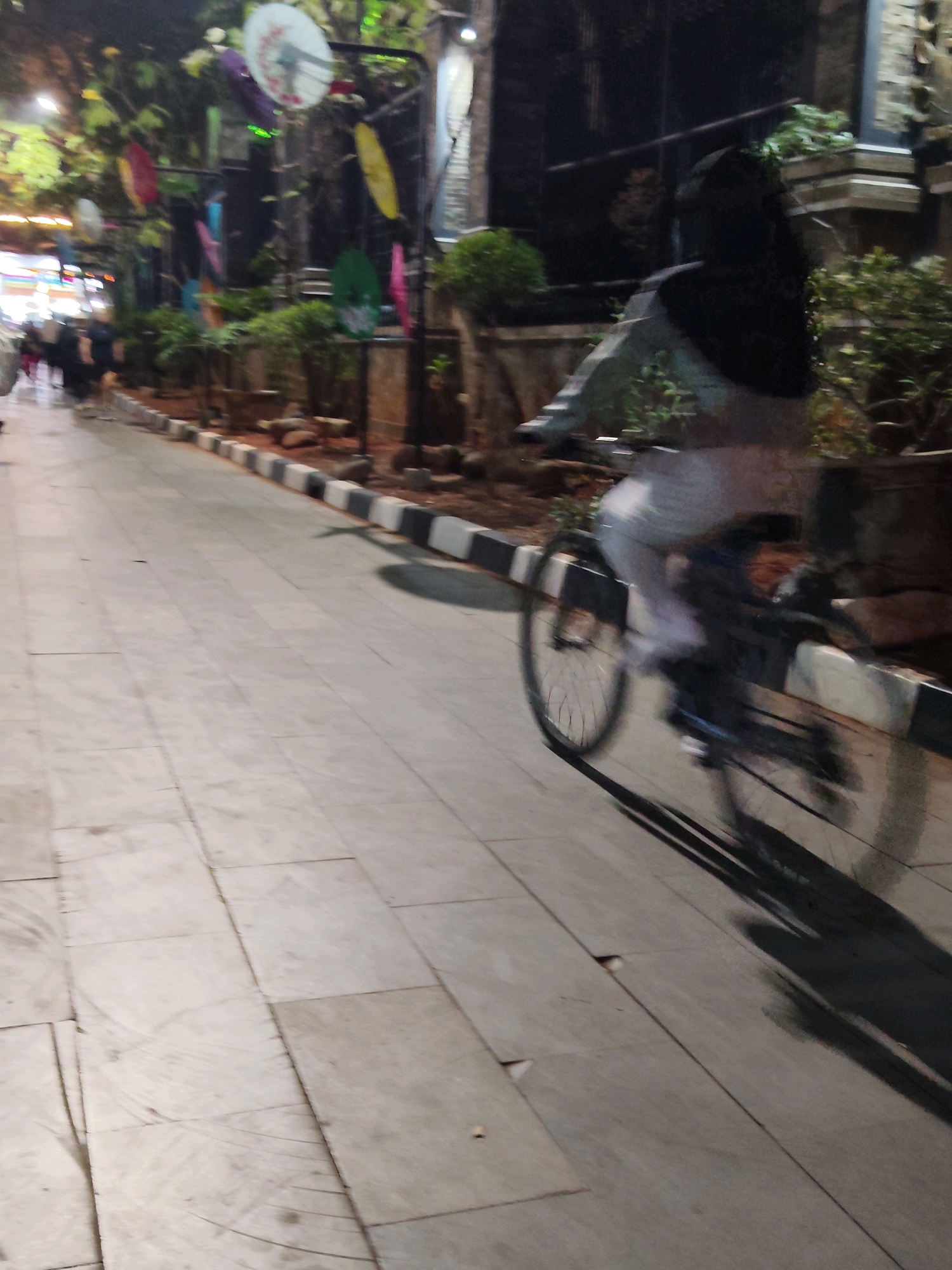 A person is riding a bicycle on a paved pathway at night in Purwakarta, Indonesia. The person, wearing a dark head covering and light-colored pants, is blurred due to motion. The bicycle's wheel and shadow are visible. The pathway is lined with decorative black and white curbs and landscaping featuring plants and rocks. In the background, colorful umbrellas are suspended from a structure, and other blurred figures can be seen walking on a separate path to the left. The scene suggests an outdoor public space, possibly a promenade or a pedestrian street, illuminated by artificial lights that cast long shadows.