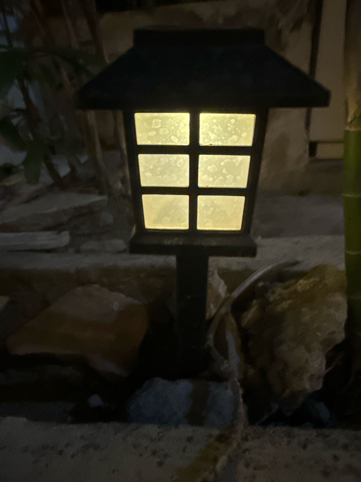 In the city of Unknown, Unknown, a traditional Japanese-style solar lantern illuminates the dusk-lit garden. The lantern, made of dark, weathered material, features a multi-paneled glass front that glows with a warm, yellowish light. The glass is speckled with what appears to be dirt or water spots. Surrounding the lantern, rough, natural stones of varying sizes create a rustic landscape. In the background, lush green foliage and stalks of bamboo suggest a tranquil outdoor setting. A long, slender lizard is visible, perched on a rock to the right of the lantern, its body slightly curved as it rests. The scene is dimly lit, hinting at the transition from day to night, with shadows cast by the surrounding elements.