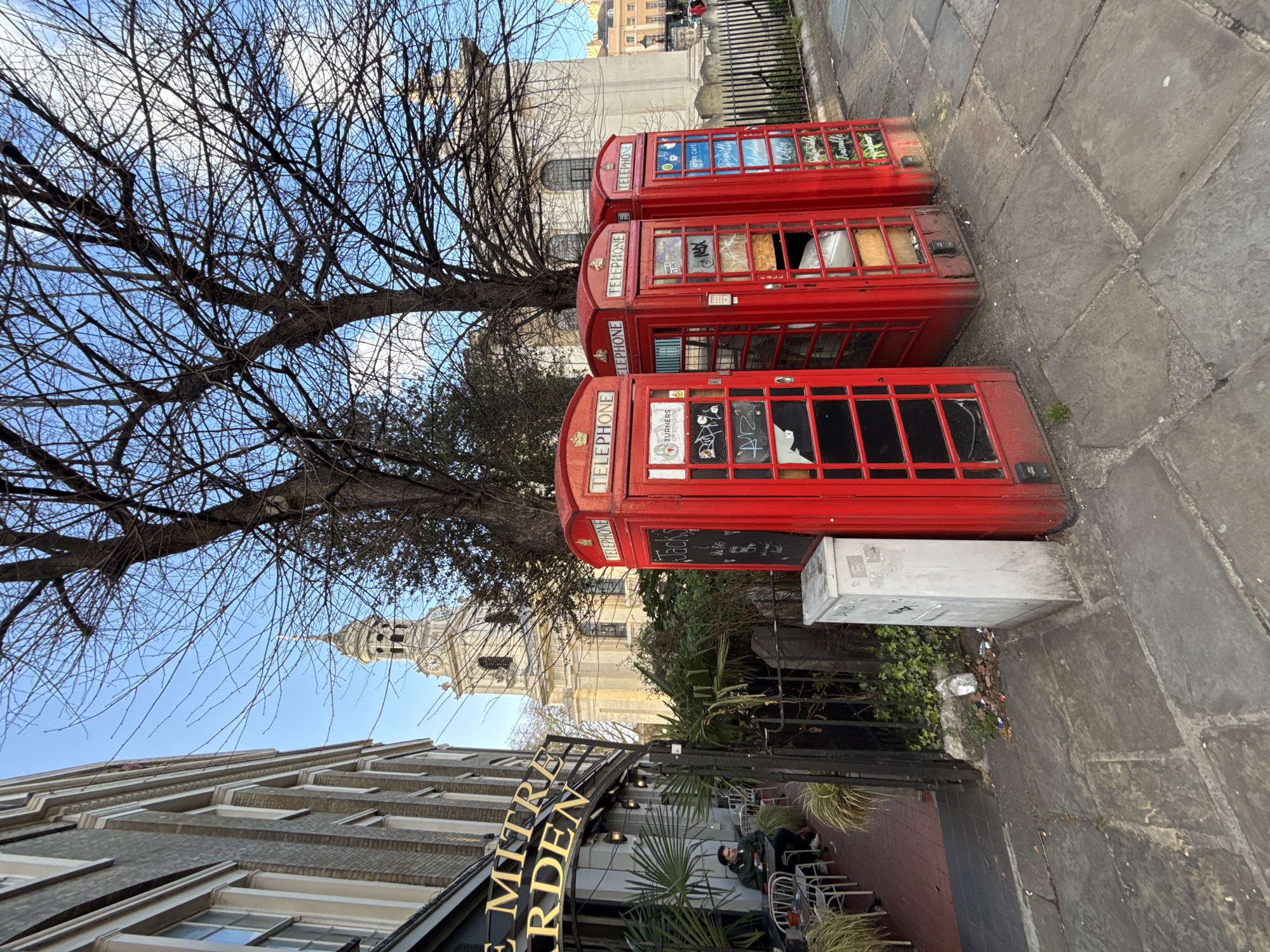 Three red telephone boxes stand in a row on a paved surface in London, United Kingdom. The boxes have graffiti and damaged windows. A bare tree with many branches looms overhead. A building with a clock tower is visible in the background. In the foreground, part of a building with the sign "THE MITRE GARDEN" is seen, with people sitting at tables outside.