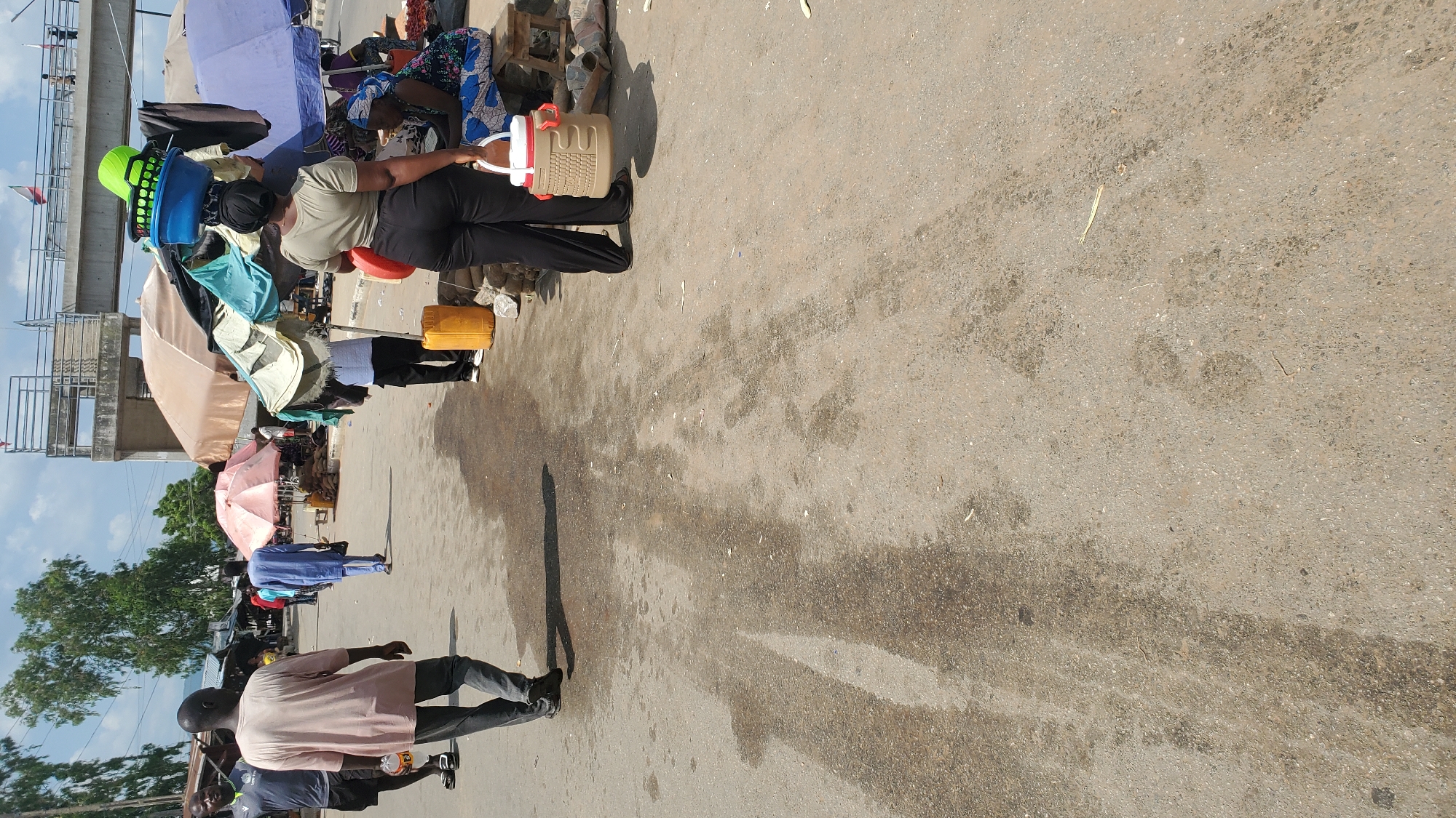 In Jalingo, Nigeria, a street market scene unfolds under a bright, partly cloudy sky. In the foreground, a person in dark pants and a light green shirt carries a tan and red insulated container with a white lid. To the left, another individual in a light pink shirt and dark pants walks away from the camera, holding a clear plastic bottle. Several market stalls are visible in the background, some shaded by umbrellas, with produce like yams displayed. People are seen walking and standing amongst the stalls. Overhead, a concrete overpass spans the area, with people visible on it. The ground is a dusty, asphalt surface, showing tire tracks and damp patches.