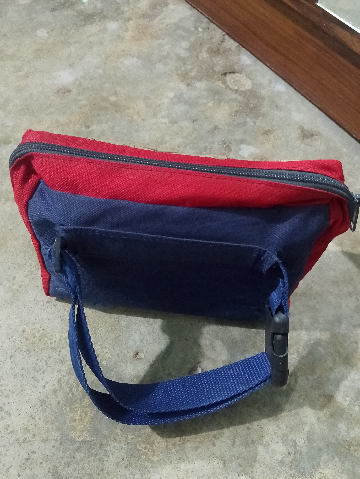 The image shows a red and blue fanny pack lying on a grey, textured surface that appears to be concrete or cobblestone. The fanny pack is oriented horizontally, with the red portion on top and the blue portion below. A black zipper runs along the top edge of the red section. A blue strap with a black plastic buckle is attached to the blue section of the fanny pack and extends outwards, creating a loop. The background is a blurred, grey, textured surface with hints of brown wood in the upper right corner, suggesting it might be a wooden piece of furniture or a structure. There is no indication of the city of Serbelawan, Indonesia, or any people present in the image.
