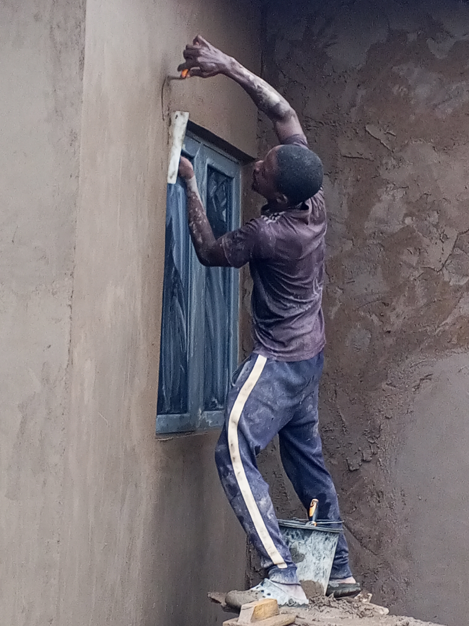 An adult male is depicted actively plastering or rendering the exterior wall of a building in Zango-ndaji, Nigeria. He stands on a makeshift platform, possibly a wooden plank or block, positioned on uneven ground. The man has dark skin and short hair, and is dressed in a dark, short-sleeved shirt and dark blue track pants with a white stripe, both noticeably stained with the wet building material.
He is focused on applying a wet, earthy-colored plaster or mud mixture to the wall around a dark-framed window using a small, orange-handled trowel held in his raised right hand. His left hand is near the window frame. The window's interior is covered with what appears to be a protective plastic sheet. Near his feet, a black bucket, splattered with the same material, holds a yellow-handled tool.
The wall exhibits areas of freshly applied, wet plaster and sections that appear to be already drying, indicating ongoing construction or finishing work. The scene is illuminated by daylight, suggesting a soft, diffused light without harsh shadows. No text is visible in the image.