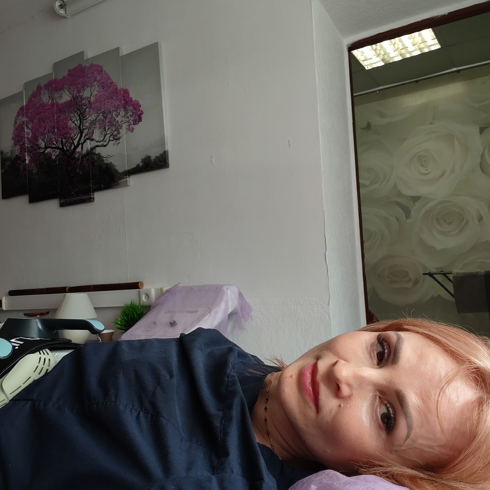 A woman with light reddish-blonde hair, fair skin, and wearing a dark blue shirt lies on a purple surface. Her face is turned towards the camera.
Behind her, on the wall, hangs a five-panel art piece depicting a tree with pink foliage against a grayscale background. Below the artwork, a bamboo rod sits above a shelf holding a white lamp and a small green plant.
To the right of the woman, a mirror or a reflective surface shows a floral pattern wallpaper and part of a light fixture on the ceiling.
The image was taken in an interior setting and the location is stated to be in the city of Chaikovsky, Russia. No specific actions or interactions are depicted beyond the woman lying down and looking at the camera.