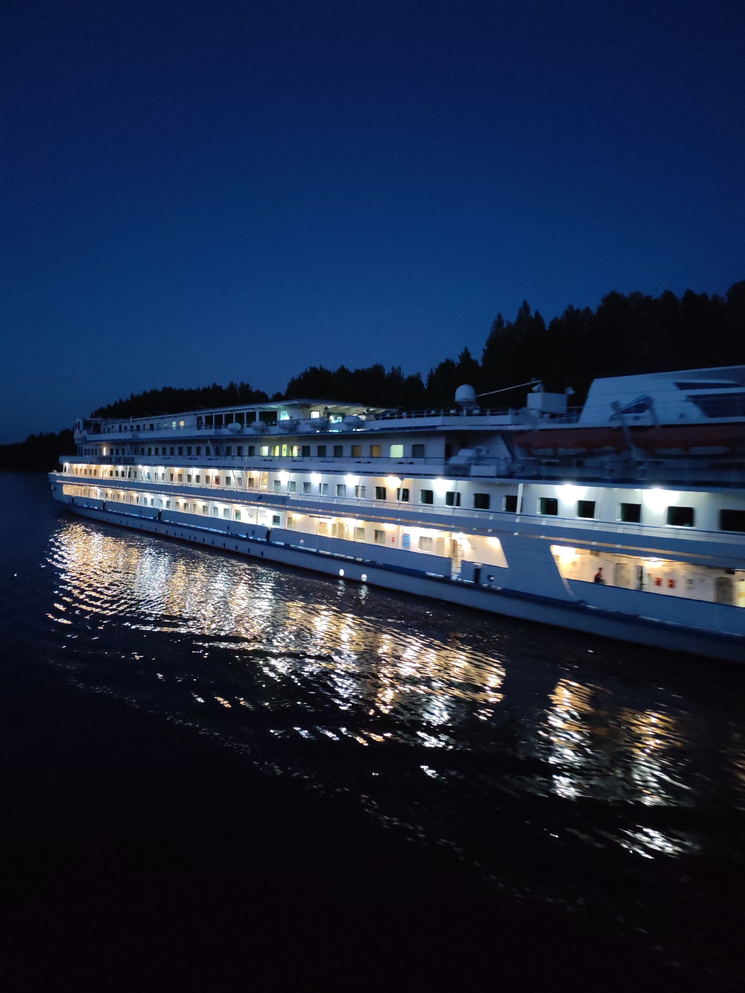 A large, white, multi-deck passenger ship is cruising on dark water at night. The ship is illuminated by numerous lights from its interior, casting bright reflections on the water's surface. The sky above is a deep, dark blue, and the silhouette of a forest can be seen on the distant shore. The scene suggests a tranquil evening journey on a body of water near a wooded area, potentially the Volga River in Depo, Russia, given the typical presence of such vessels and landscapes in the region. There are no visible people on the decks or in the water. The ship appears to be moving forward, indicated by the disturbed water.