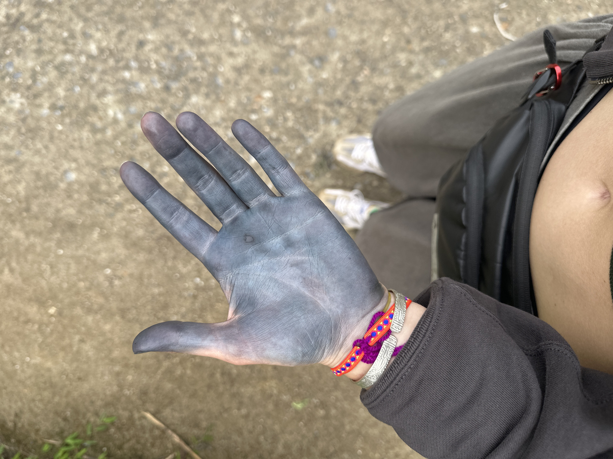 The image shows a person's hand covered in a grey-blue substance, possibly dye. The hand is held out towards the viewer. The person is wearing multiple bracelets on their wrist and a dark-colored hooded sweatshirt. A black backpack or bag is visible slung over their shoulder, with a glimpse of their skin visible under the bag strap. The person is standing on a paved or gravel surface. Based on the prompt, this is located in Ban Ho, Vietnam.
