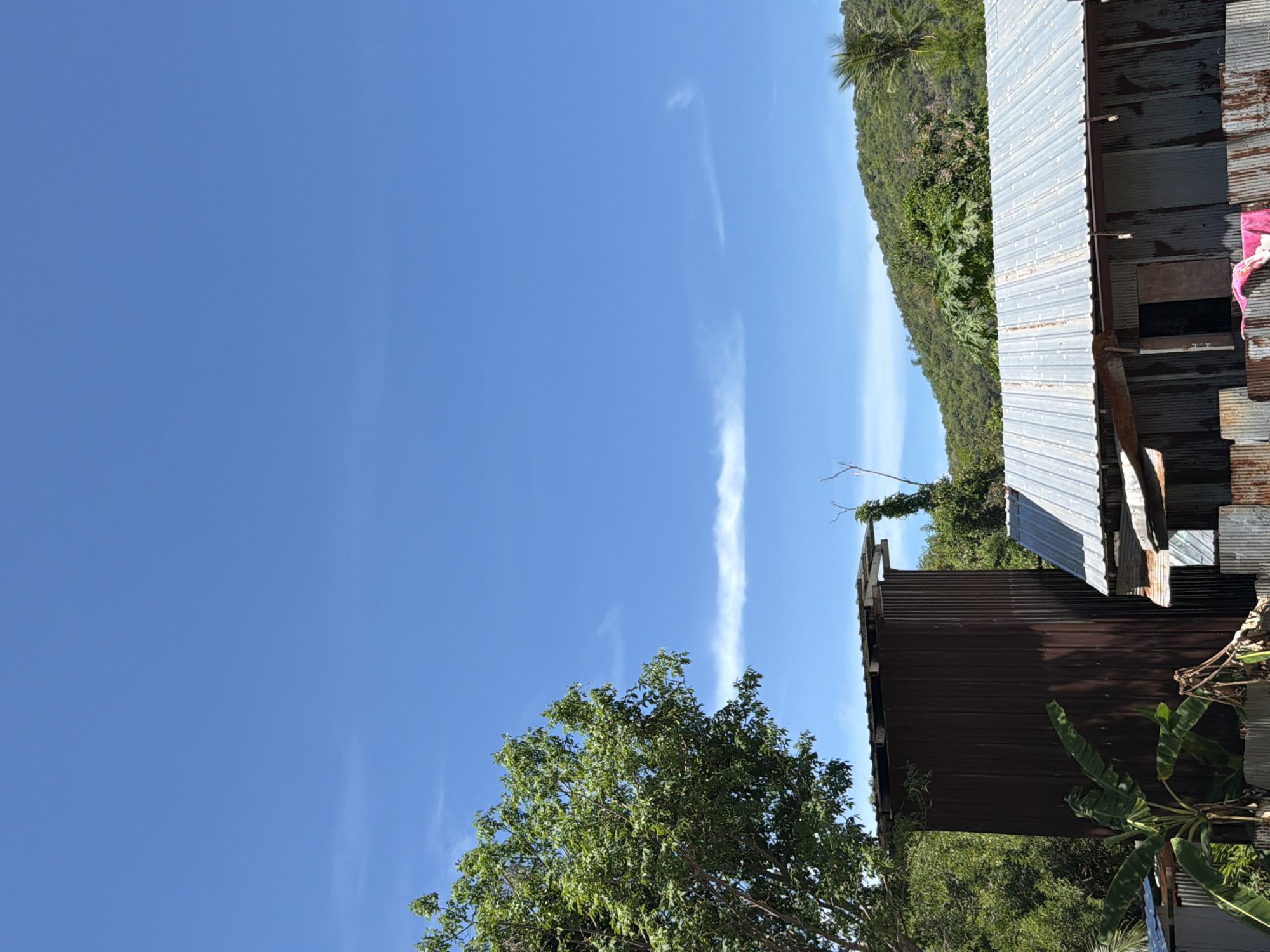 The image shows a bright, sunny day in Baan Bo Phut, Thailand, with a clear blue sky and sparse white clouds. In the foreground, there is a tall, dark brown corrugated metal structure, possibly an outbuilding or shed. To its right, a single-story building with a corrugated metal roof and walls is visible. This structure appears weathered and somewhat dilapidated, with an open doorway and a pink garment draped over its exterior. A banana tree with large leaves stands to the left of the brown structure. In the background, lush green hills rise under the expansive sky. The overall scene suggests a rural or semi-rural setting, typical of areas outside major city centers in Thailand.