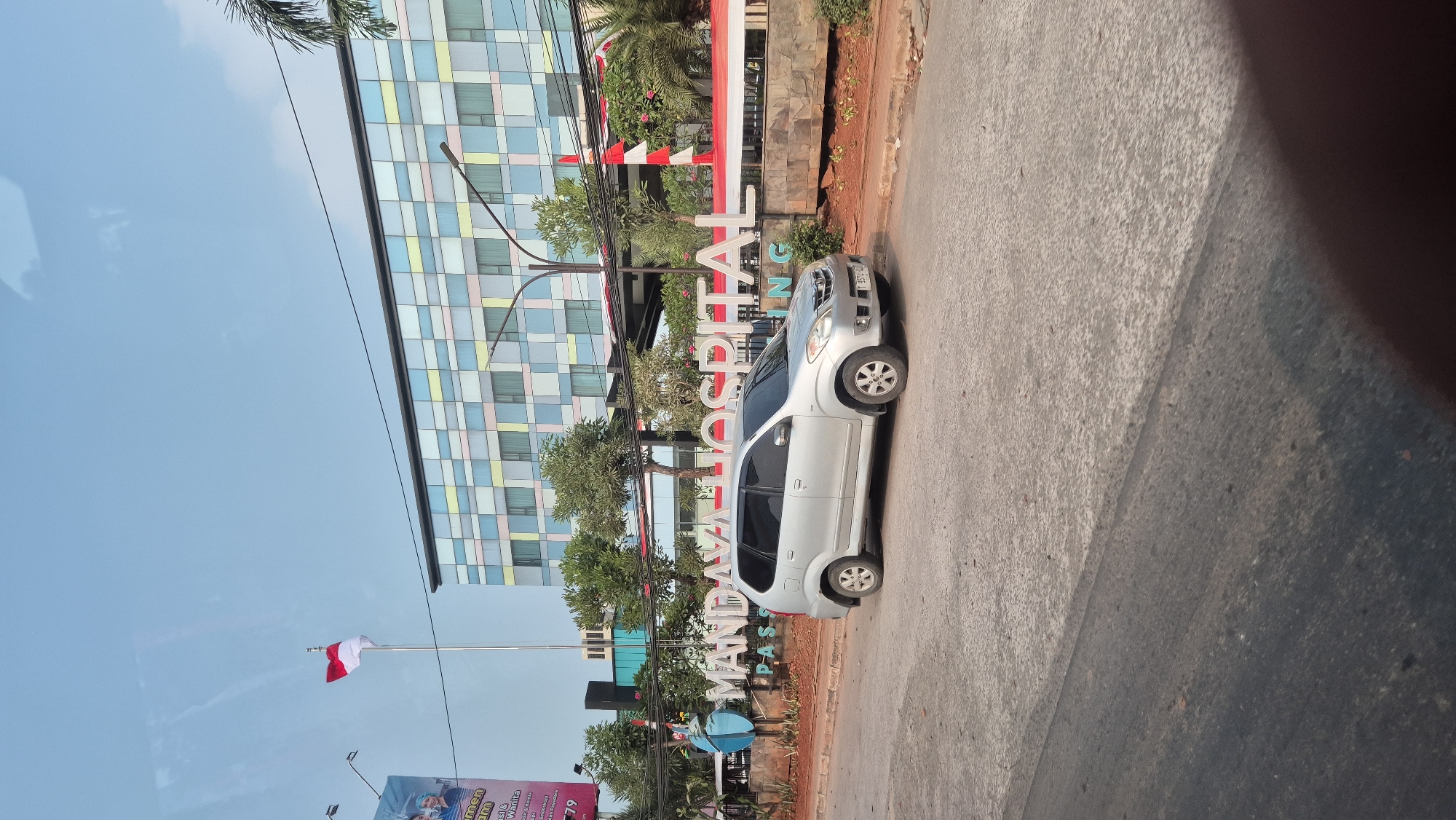 The image depicts the entrance of Mandaya Hospital in Karawang, Indonesia. A silver minivan is parked in front of the hospital's prominent signage. The building itself has a modern facade with colorful rectangular panels in shades of blue, yellow, and pink. An Indonesian flag flies from a flagpole to the left of the building, and red and white banners are displayed along the entrance. Trees and greenery surround the area, with a street in the foreground. The sky is clear and blue.