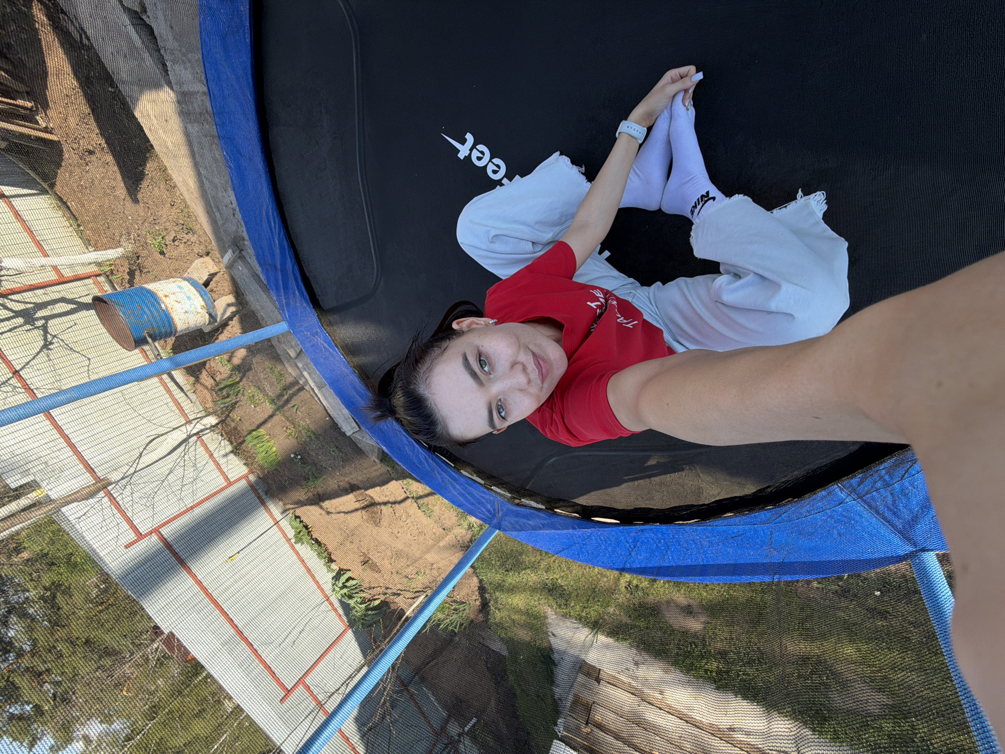 A young woman lies on a trampoline in Chaikovsky, Russia, looking at the camera. She wears a red t-shirt, light-colored pants, and white Nike socks. The trampoline has a blue safety net. A blue and white barrel sits outside the net. The background shows a fenced area with greenery.