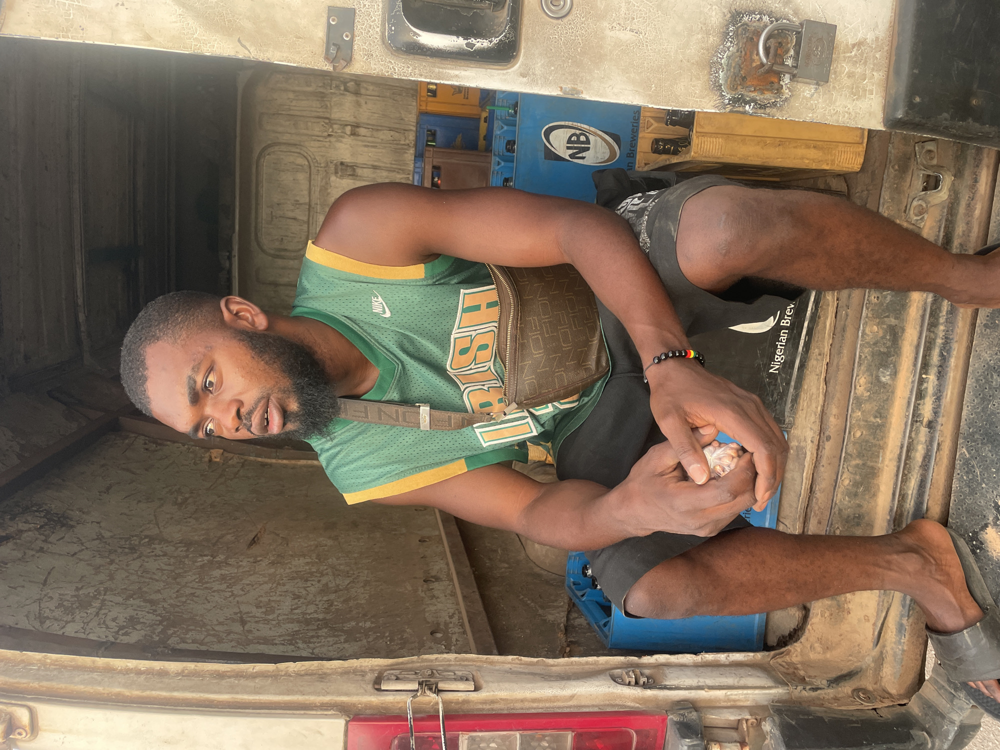 A man sits in the back of a vehicle, possibly a truck or van, in what is likely a bustling area of Jos, Nigeria. He is wearing a green and gold jersey, black shorts, and sandals. A small brown bag is slung across his chest. He holds a small bag of what appears to be food in his hands. Behind him are stacks of blue and yellow beer crates. The inside of the vehicle is worn and shows signs of use. The lighting indicates it is daytime.