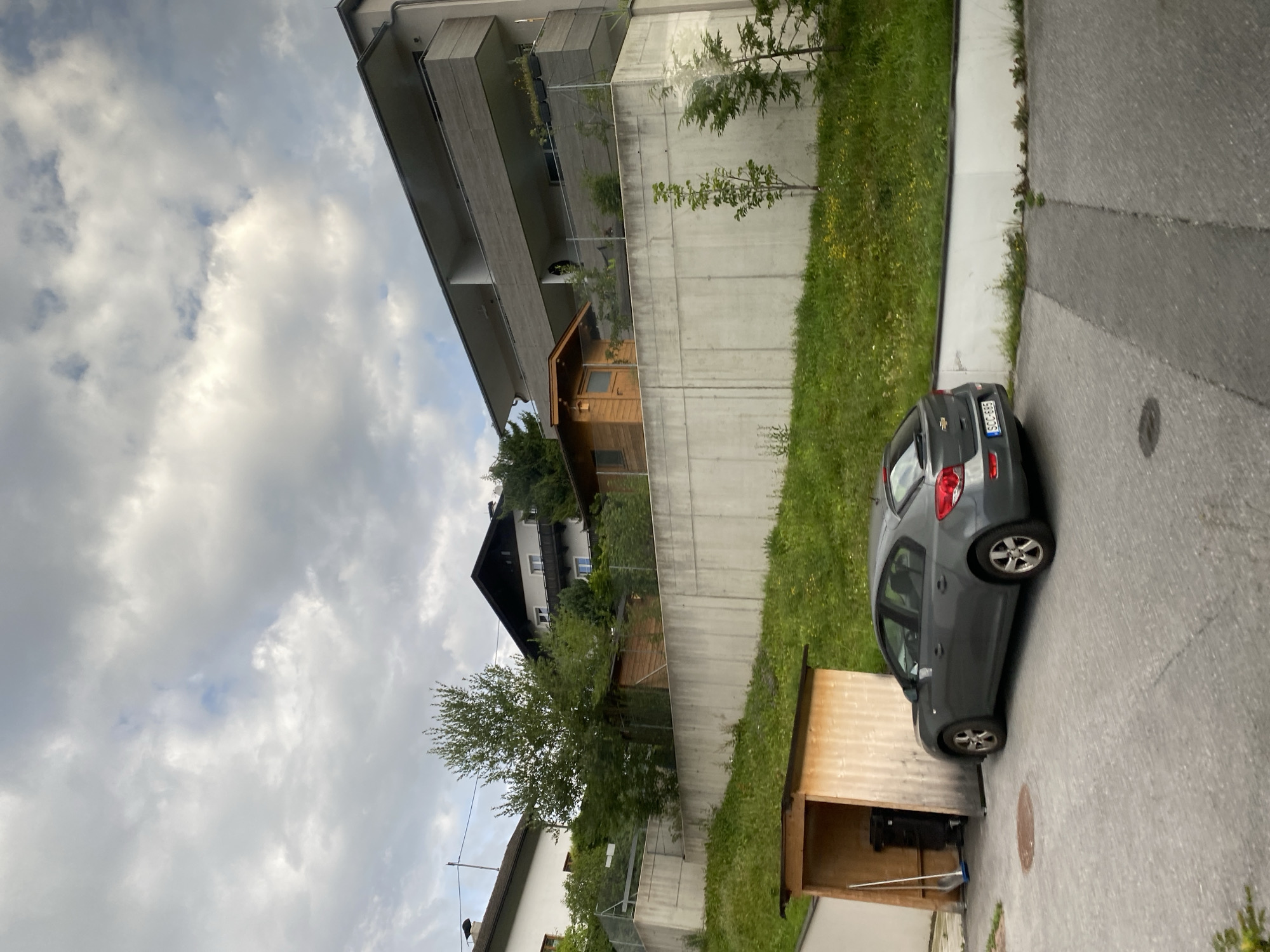 In Reith bei Seefeld, Austria, a dark grey Chevrolet Cruze sedan is parked in a paved area next to a small wooden lean-to shelter containing a black garbage bin and a broom. The car is facing away from the viewer, with its rear bumper and license plate (SCC-665) visible. Behind the car and shelter, a concrete retaining wall separates the lower paved area from a grassy hillside with scattered yellow flowers. Atop the hillside, a small wooden shed with a window and door is situated. Further up the slope, a modern multi-story building with balconies and a grey facade stands. Trees and other houses are visible in the background, with a cloudy sky overhead. The scene appears to be a residential area with a mix of older and modern architecture.