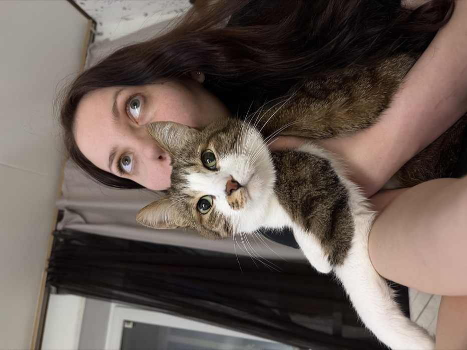 A woman with long brown hair holds a cat. The cat has a white and brown face with green eyes. The woman looks upward. She has light skin and dark eyebrows. A window with dark curtains is visible behind them. The background suggests they are in a room with a bed.
