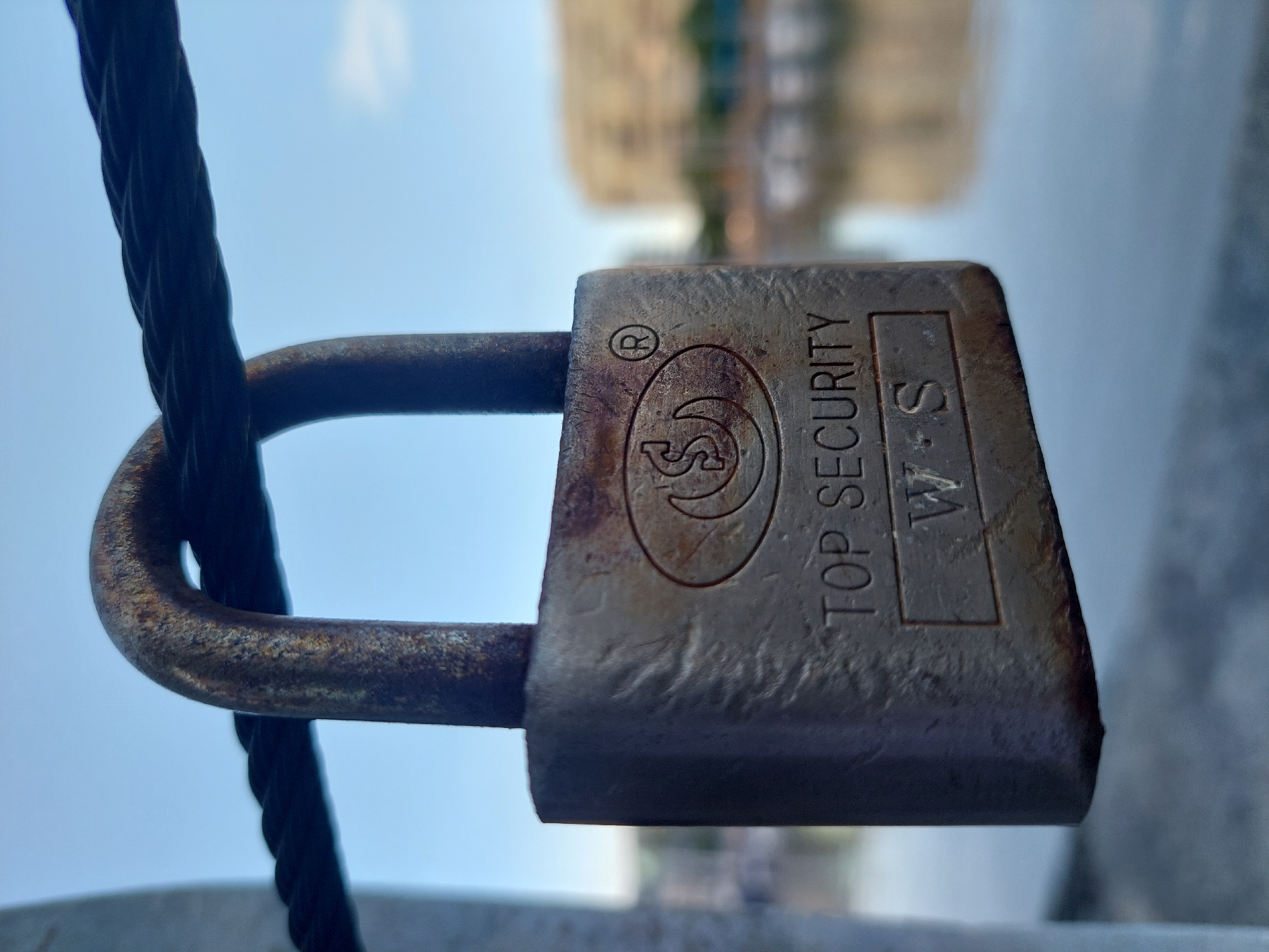 A close-up photograph shows a weathered metal padlock attached to a thick, braided wire. The lock displays the brand name "TOP SECURITY W.S" and features a circular logo. The background is blurred, but the suggestion is that it is outdoors, maybe a city scene. Location: Buenos Aires, Argentina. Likely near a bridge or other infrastructure. People not visible. Subject is a security device and wire. Objects present: Padlock, wire. Actions/interactions: Padlock is attached to the wire, perhaps securing something. Environment: Urban, possibly near infrastructure.