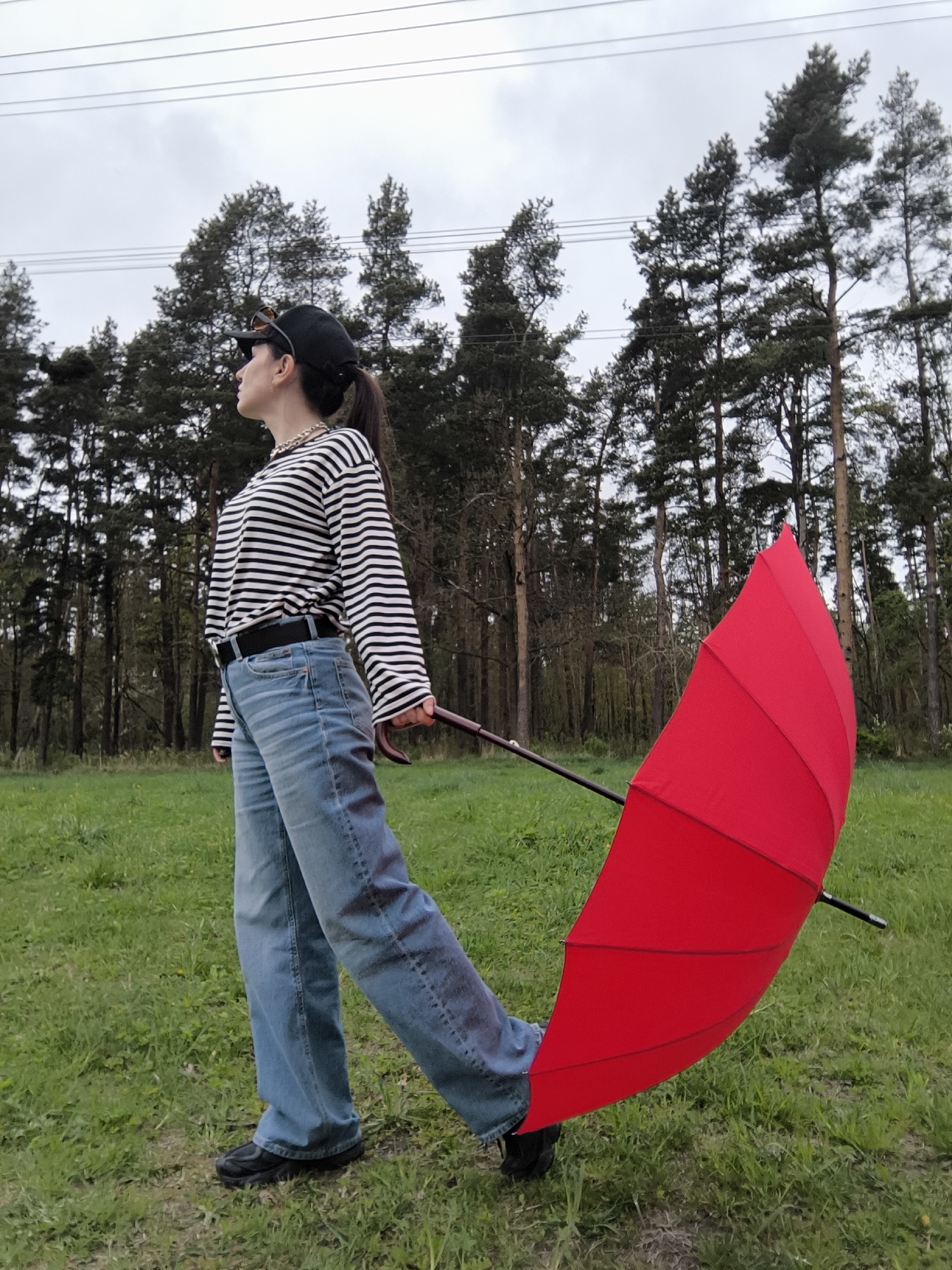A woman stands in a grassy field near a forest, holding a red umbrella. She is wearing a black and white striped shirt, blue jeans, and a black cap, with a necklace visible at her neck. The woman is looking to her left, away from the camera. Power lines are visible in the sky above the trees. The location is a grassy area next to a dense forest, with an overcast sky.