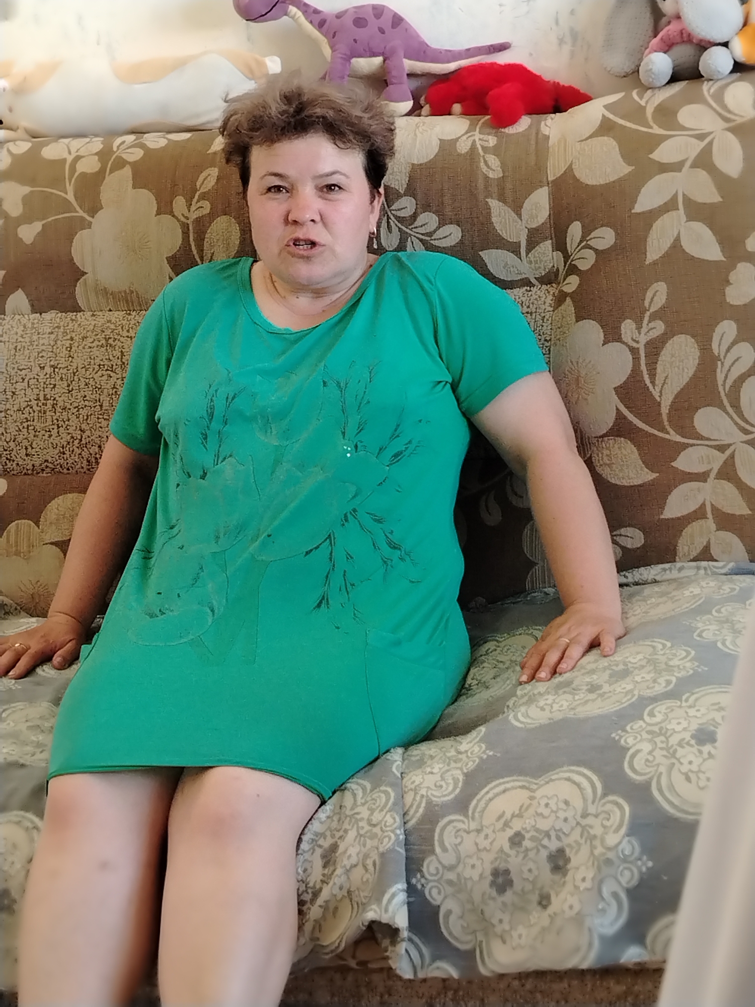 A woman is sitting on a floral patterned sofa. She wears a green dress with a darker green tree-like design on the front. Her hair is short and curly. She is looking directly at the camera and speaking. Behind her on the sofa are several stuffed toys, including a purple dinosaur and a red plush toy. The image was taken in Noviy, Russia.