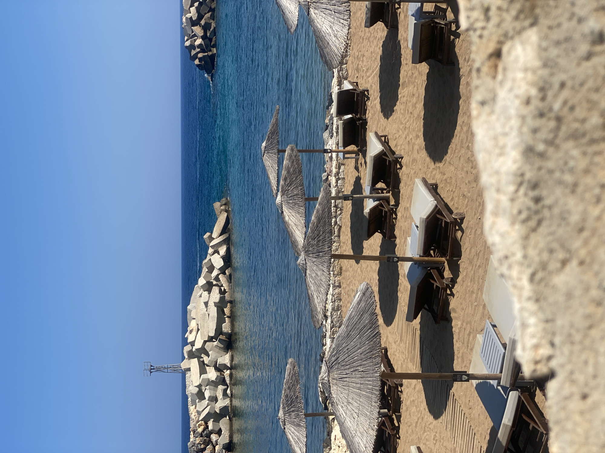 The image shows a beach scene in Panormos, Greece, on a clear, sunny day. The foreground is dominated by a sandy beach with rows of empty lounge chairs and straw umbrellas. Wooden walkways are laid out on the sand. In the background, a calm, deep blue sea meets a clear blue sky. The sea is partially enclosed by large, grey concrete breakwaters that form a harbor or protected swimming area. A tall, metallic structure with a railing, possibly a beacon or a small observation post, stands on one of the breakwaters. The overall impression is one of a serene and well-maintained beach resort.