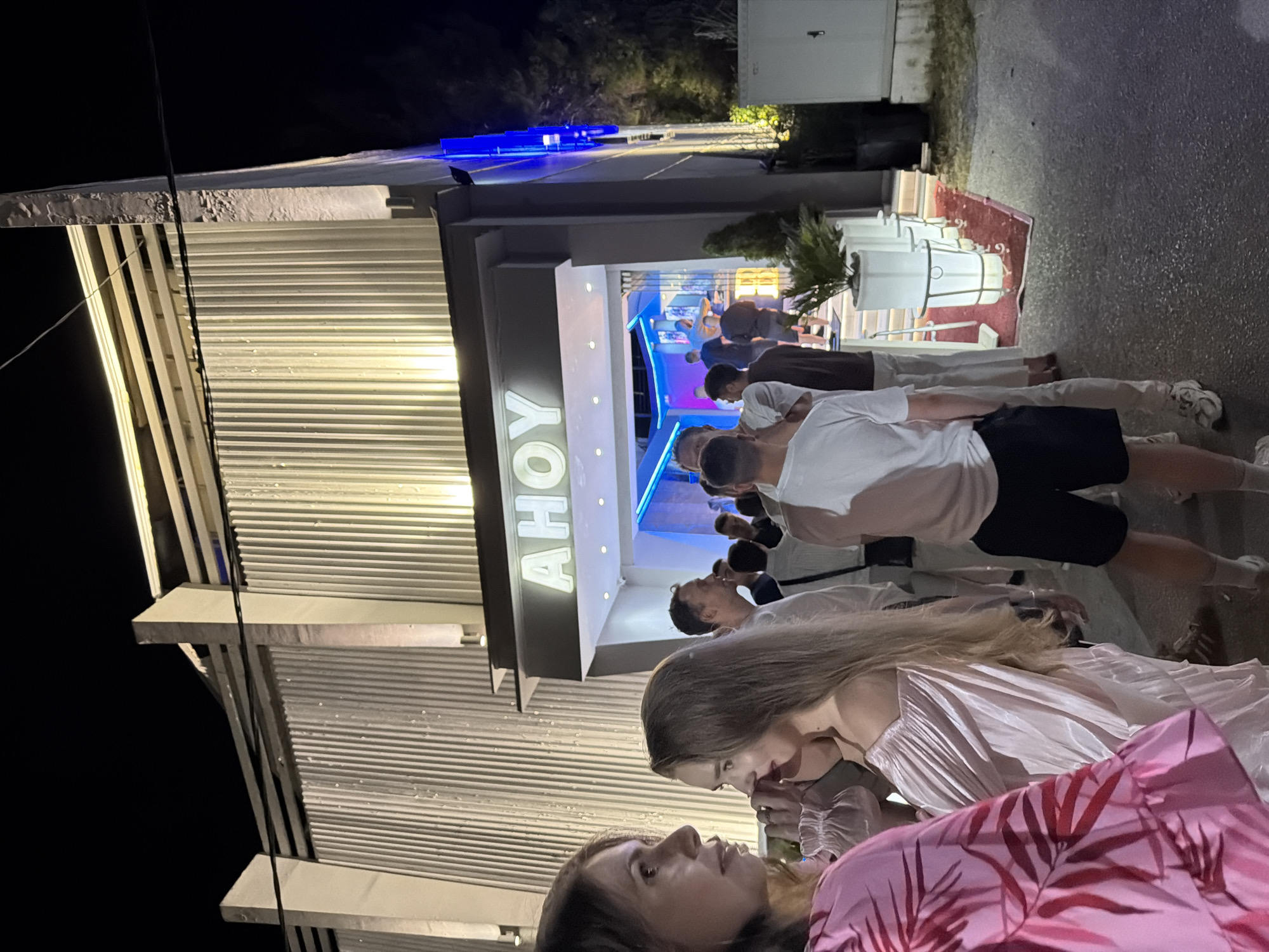 The image captures a nighttime scene outside a venue labeled "AHOY" in Kassandra Municipal Unit, Greece. A group of people are gathered in front of the entrance, which is illuminated by bright lights. Some individuals are facing the camera, while others are interacting with each other or looking towards the entrance. The architecture of the building features a ribbed facade and prominent concrete pillars. The ground appears to be asphalt or a similar paved surface. The overall atmosphere suggests a social gathering or event taking place at the establishment.